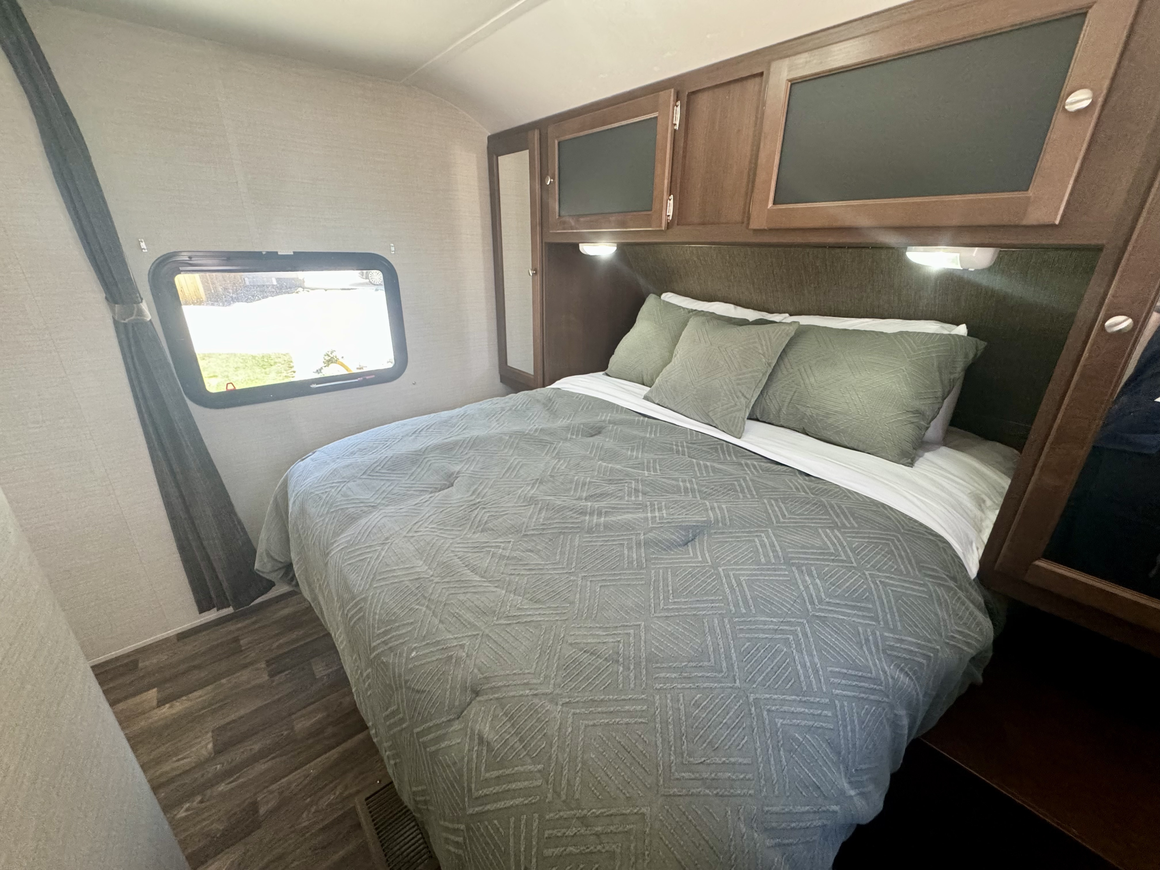 RV rental bedroom showcasing a bed with gray bedding and green pillows, wood overhead cabinets, and a window with a curtain.. Keystone RV Bullet Ultra Lite 2015