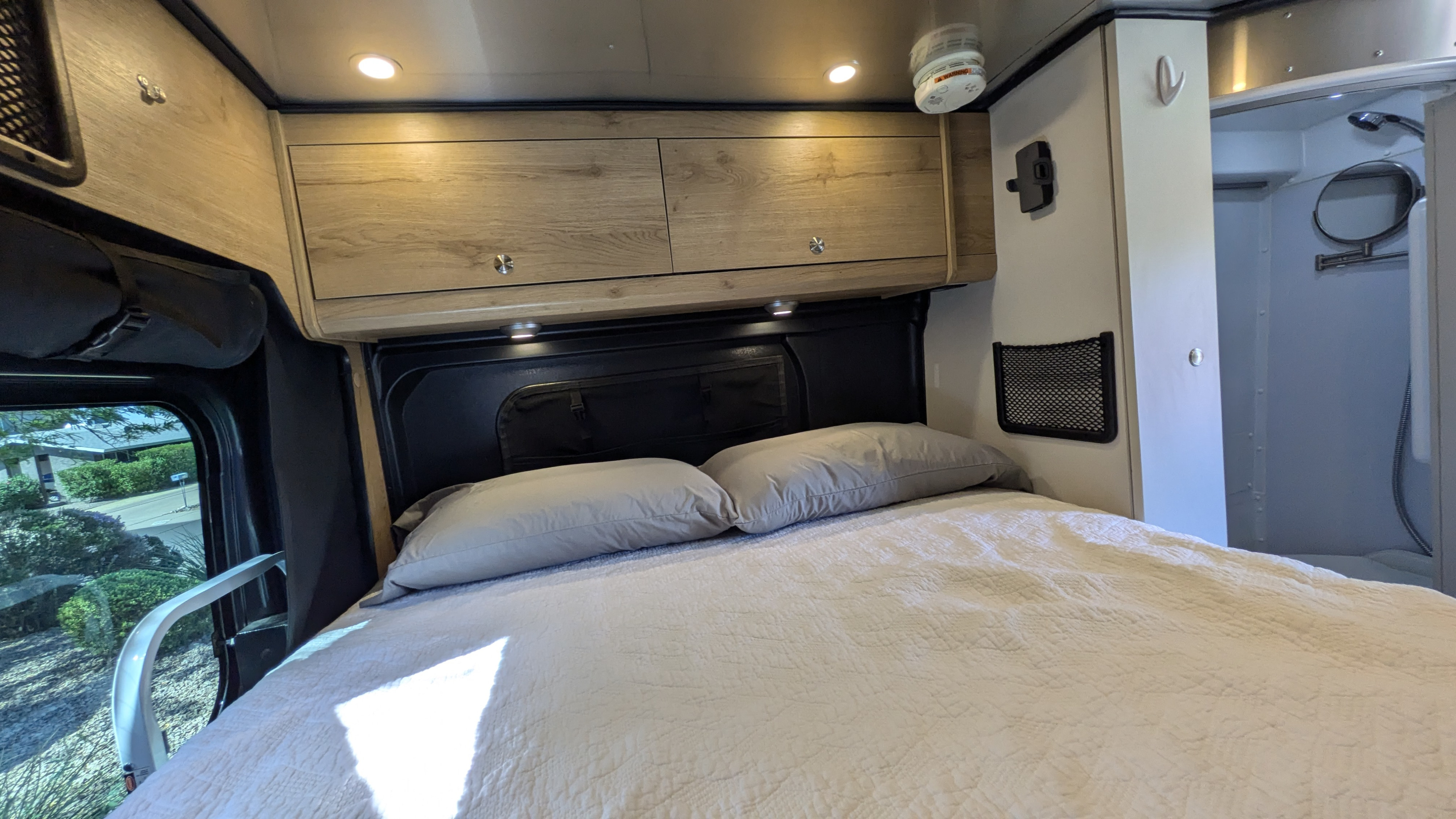 View inside an RV rental showing a comfortable bed with pillows, wood-grain overhead cabinets, and a compact shower stall.. Airstream Rangeline 2023