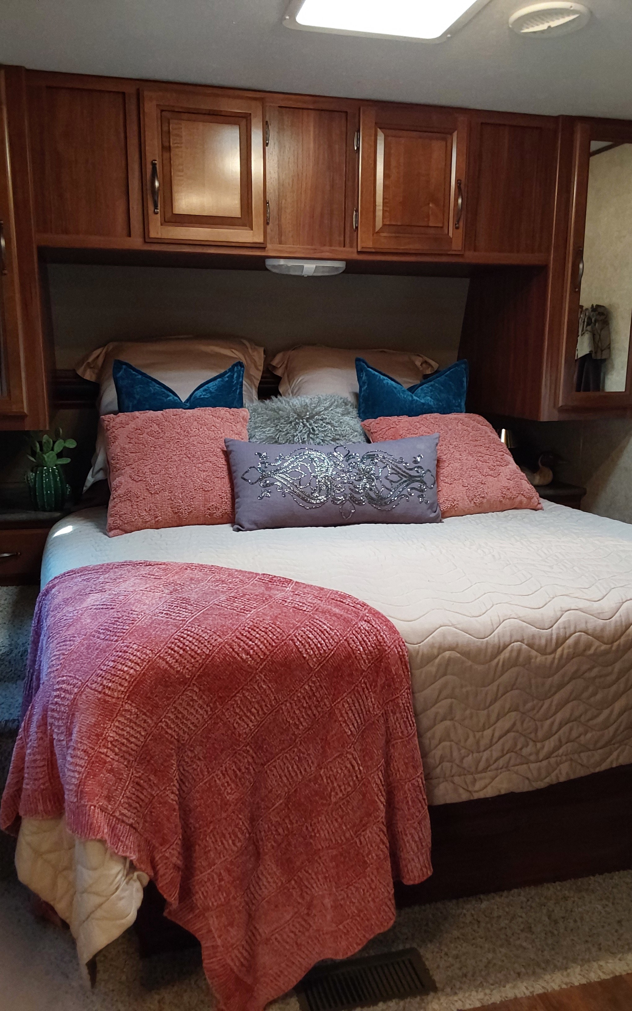A cozy RV rental bedroom: bed with vibrant pillows (teal, pink, grey, lavender) & a red blanket, surrounded by warm wood cabinets.. Keystone RV Cougar 2015