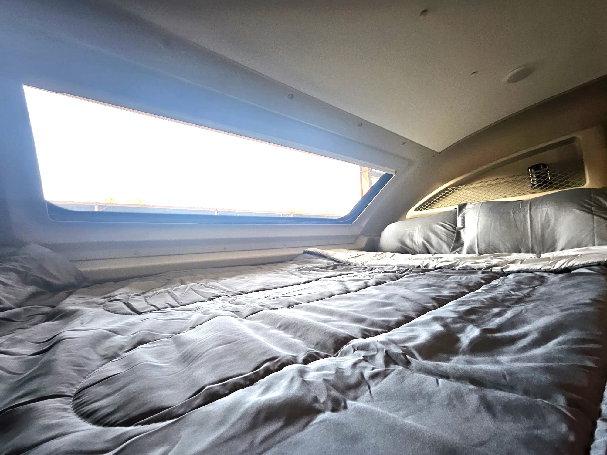 Interior of an RV rental: a made bed with gray comforter and pillows, beside a wide, bright window. Cozy sleep space.. Jayco Redhawk 2024