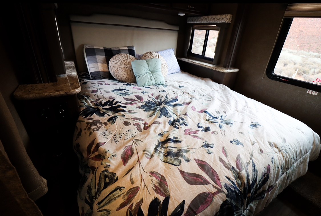 RV rental interior: a comfy bedroom with floral bed, pillows, and a window overlooking a desert landscape.. Thor Freedom Elite 2019
