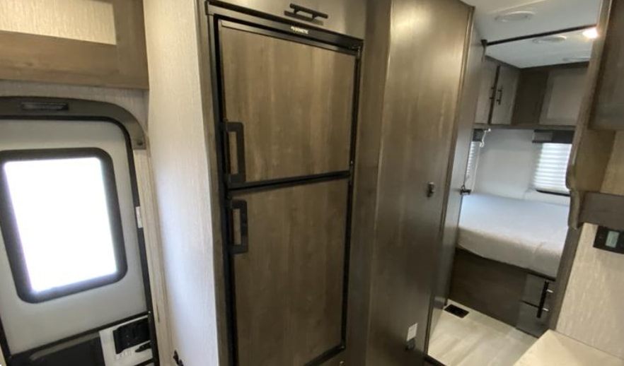 Inside an RV rental: a compact refrigerator, main entry door, and a cozy bedroom with bed and cabinets.. Forest River Coachmen Leprechaun 2023
