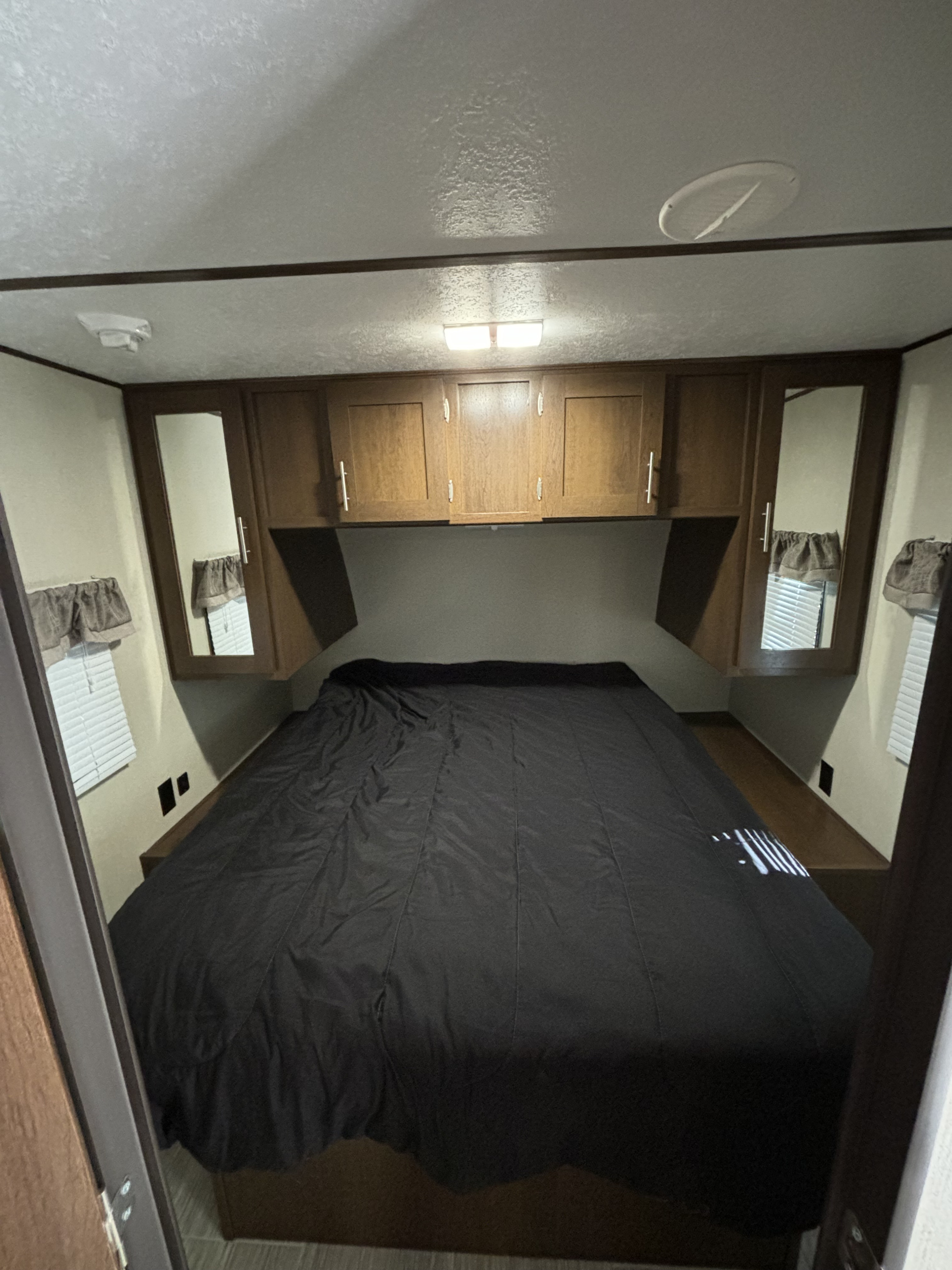 Cozy RV rental bedroom featuring a dark bed, wooden overhead cabinets, and windows with blinds.. Avenger ATI 2019