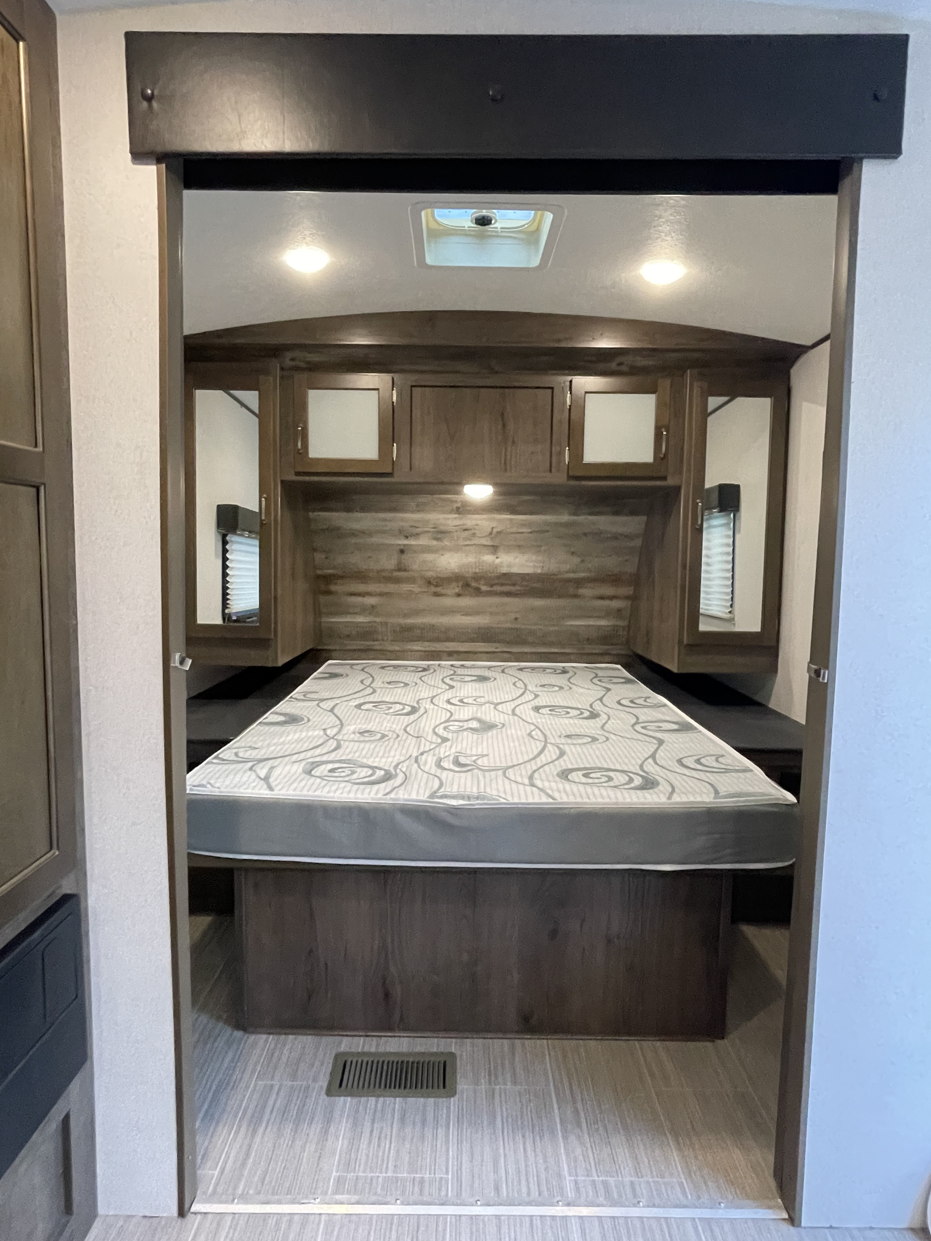 Modern RV rental bedroom: comfy bed, wood headboard, mirrored overhead storage, and bright interior lighting.. Keystone RV Passport Grand Touring 2019