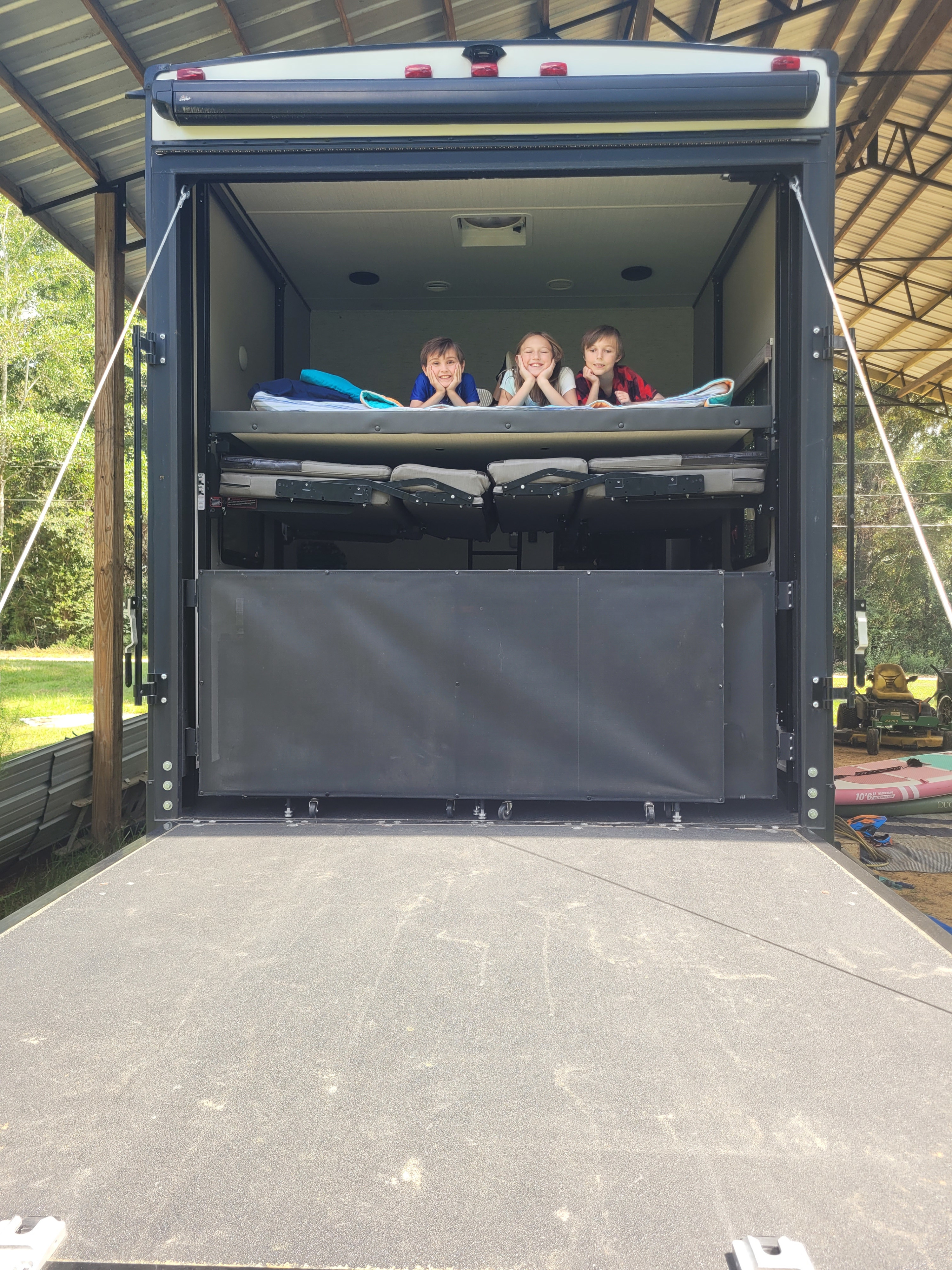 Three smiling kids inside an RV rental toy hauler, enjoying the bunk beds and open rear ramp, ideal for family trips.. Grand Design Momentum 2020
