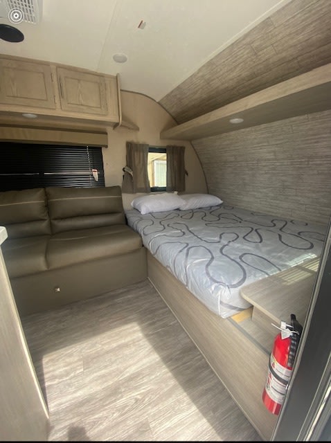 Cozy RV rental interior featuring a comfortable bed with patterned sheets, a brown sofa, and overhead storage.. Forest River Aurora Aurora Light 2023