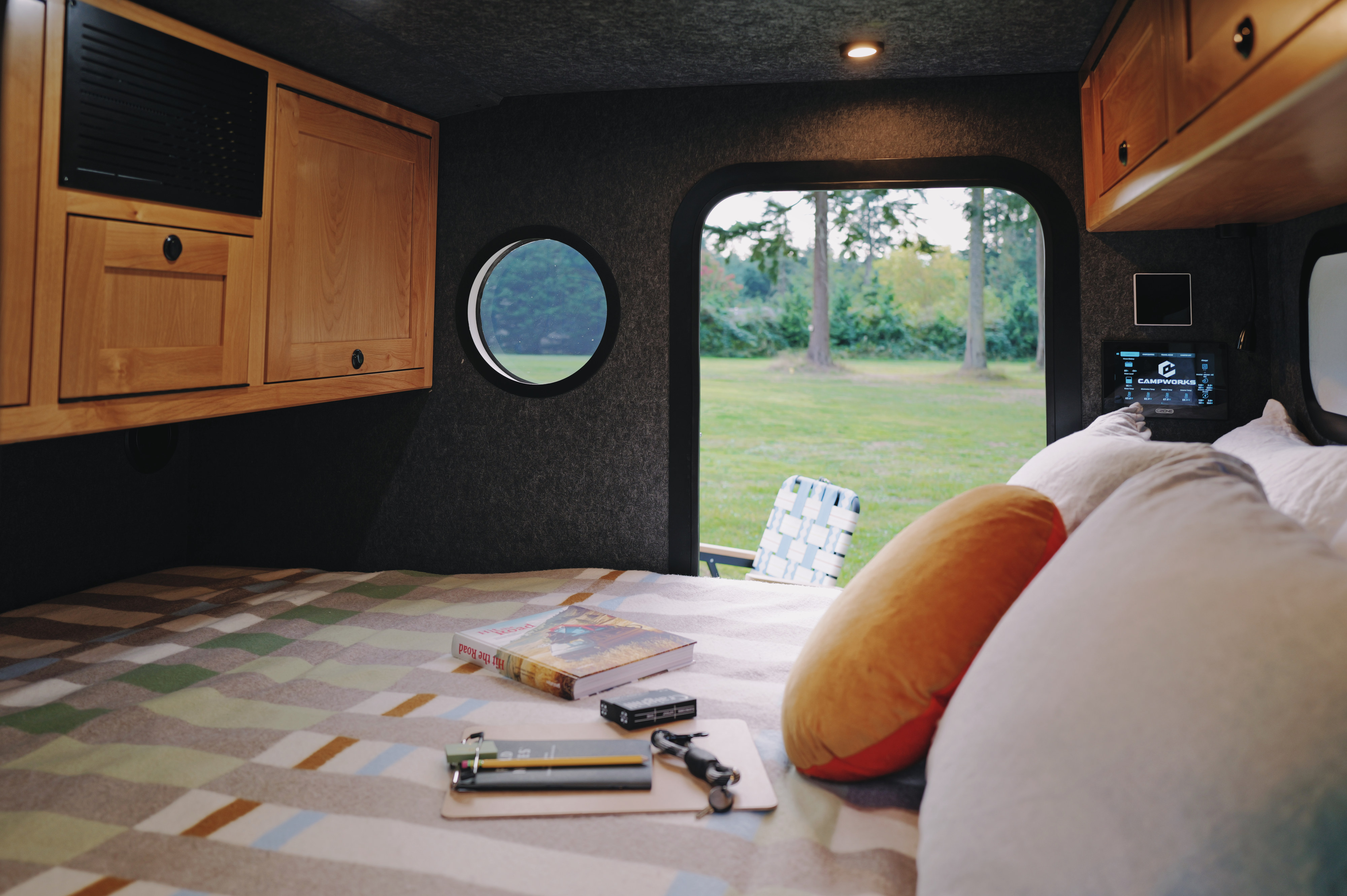 Cozy RV rental interior features a patterned bed, wood cabinets, a tech control panel, and a scenic outdoor view.. Campworks NS-1 Electrified Pro 2024
