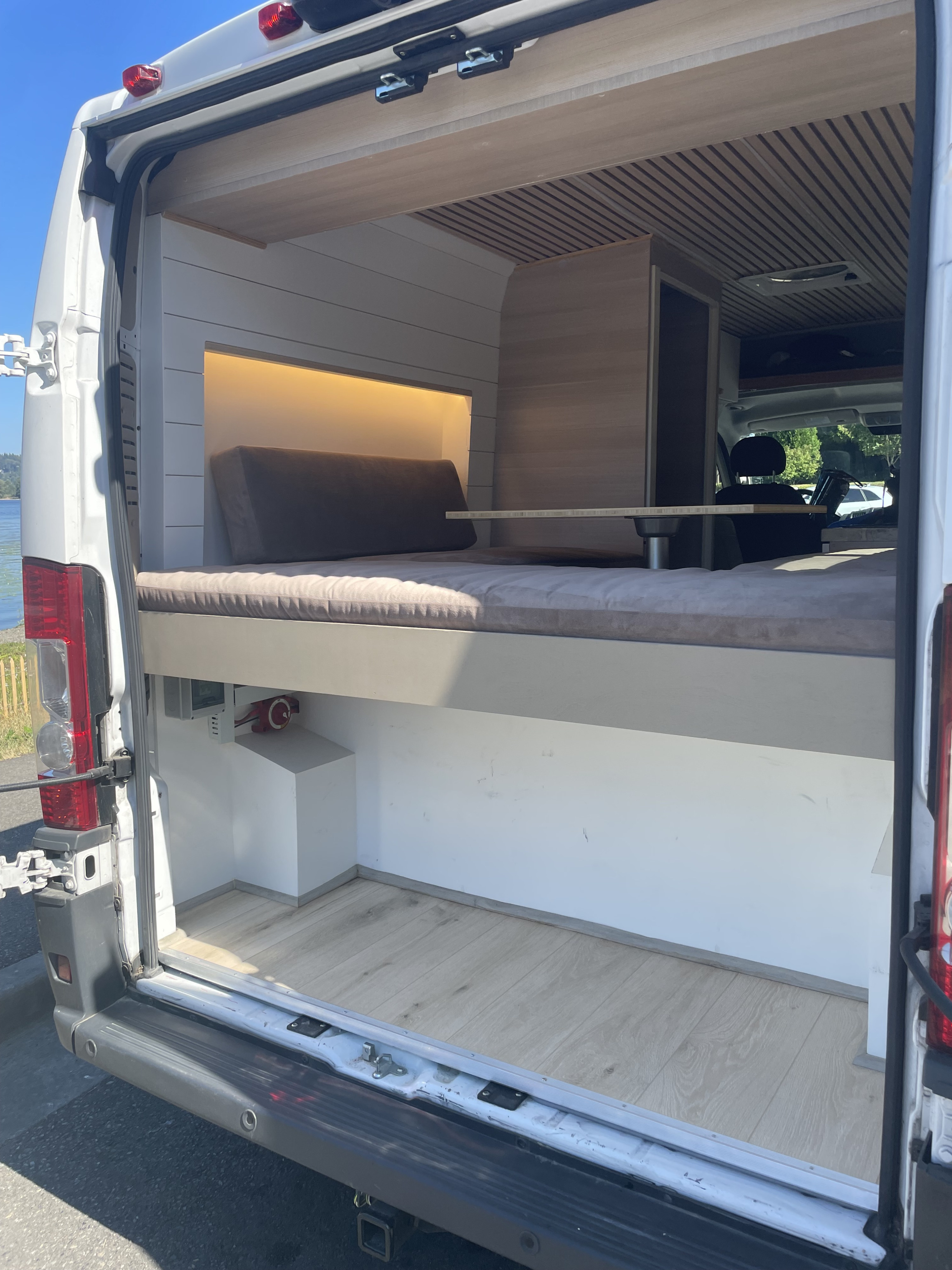 Step into this modern RV rental's customized interior, featuring a comfy bed, compact table, and cozy wood accents.. Vanberry Promaster 2018