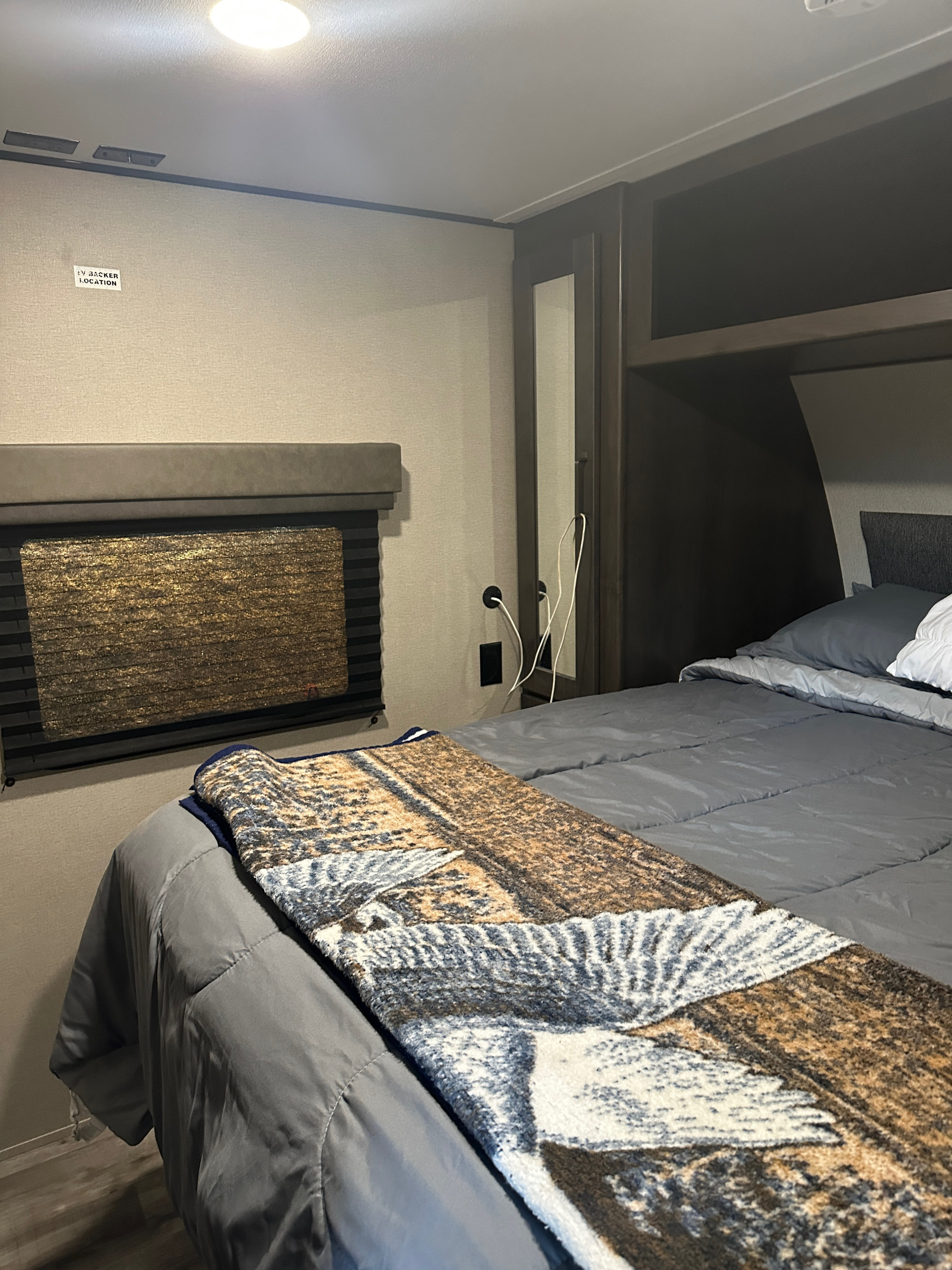 Inside an RV rental, a comfy bedroom features a grey bed with a patterned blanket, built-in storage, and a wall prepped for a TV.. Grand Design Transcend Xplor 2023