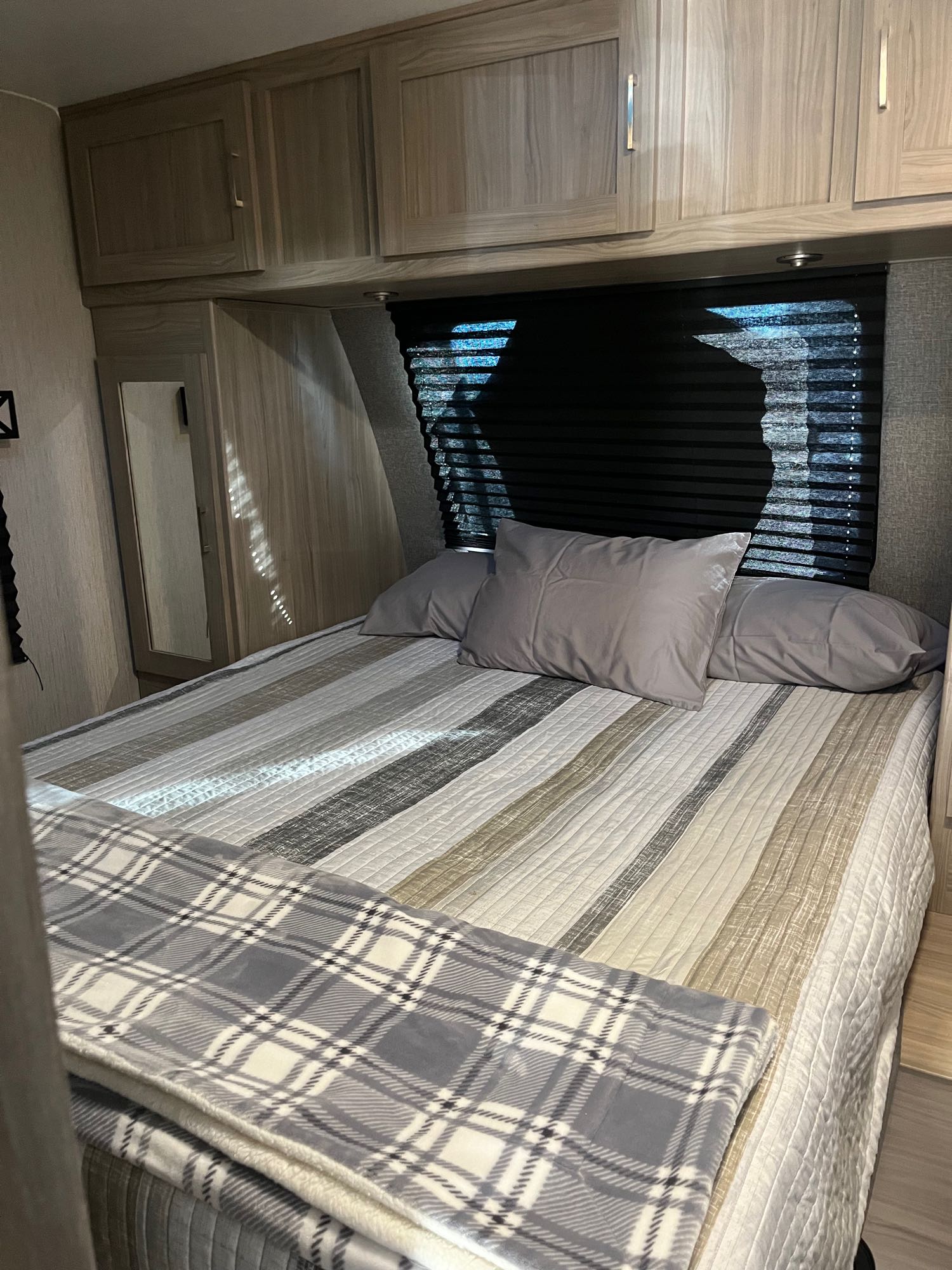 Inside a compact RV rental bedroom: bed with grey pillows, striped comforter, plaid blanket, and light wood cabinetry.. Coachmen Freedom Express Ultra Light 2020