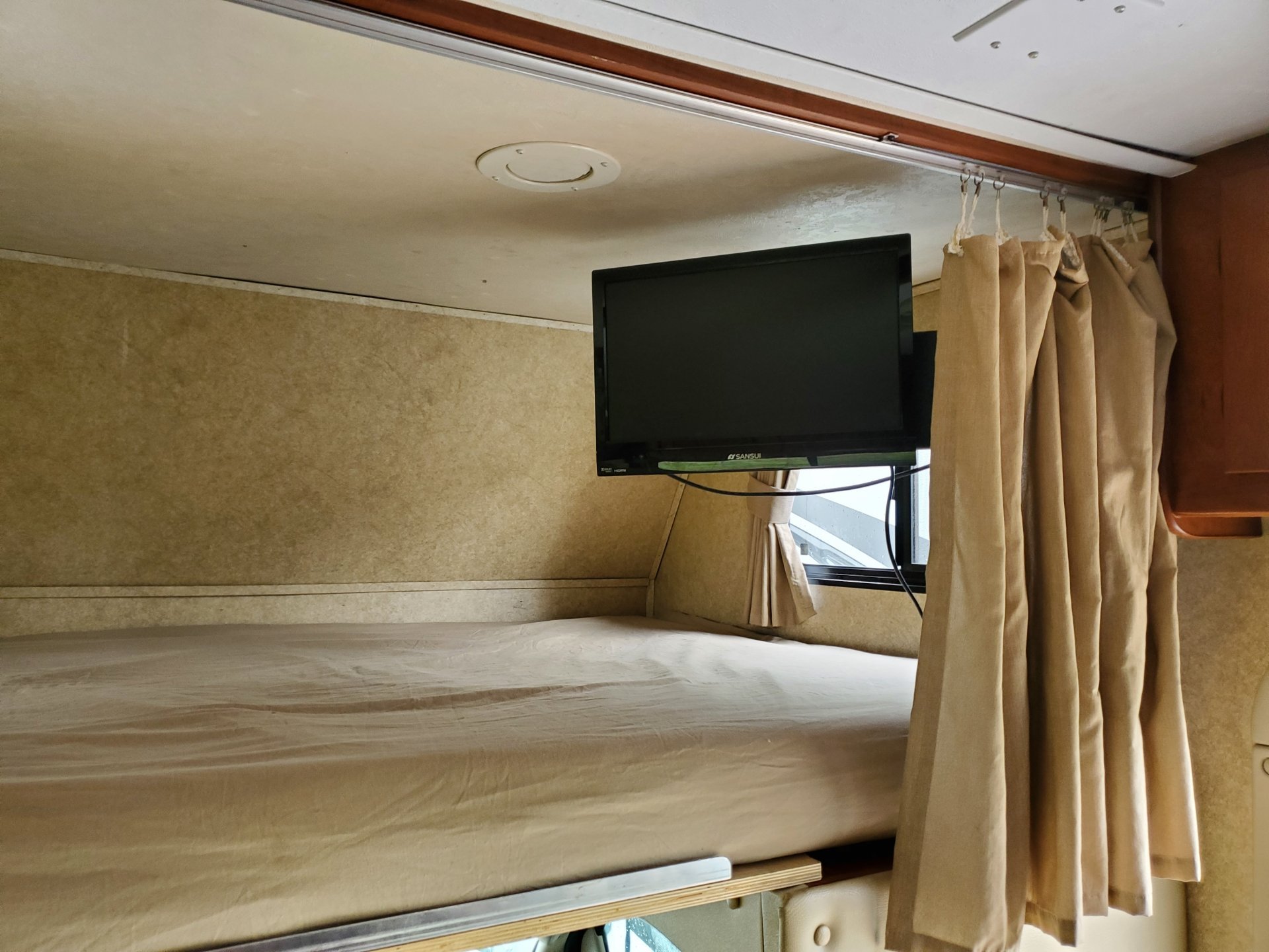 RV rental interior: cozy upper bunk bed, wall-mounted SANSUI TV, and light-colored privacy curtains enhance comfort.. Forest River Sunseeker LE 2012