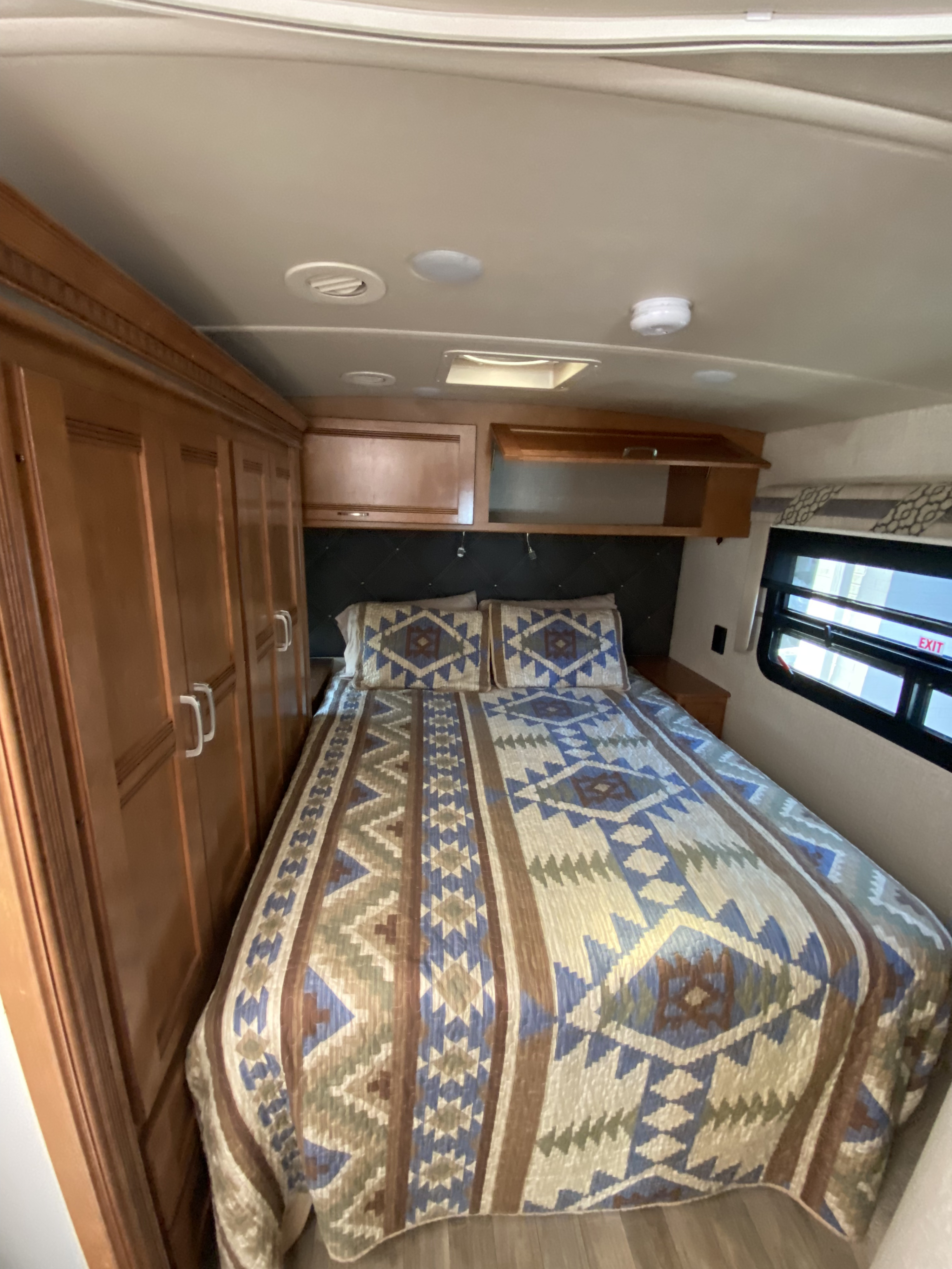 Step inside this cozy RV rental bedroom featuring a vibrant Southwestern quilt, wood cabinetry, and a bright window.. Winnebago Aspect 2019