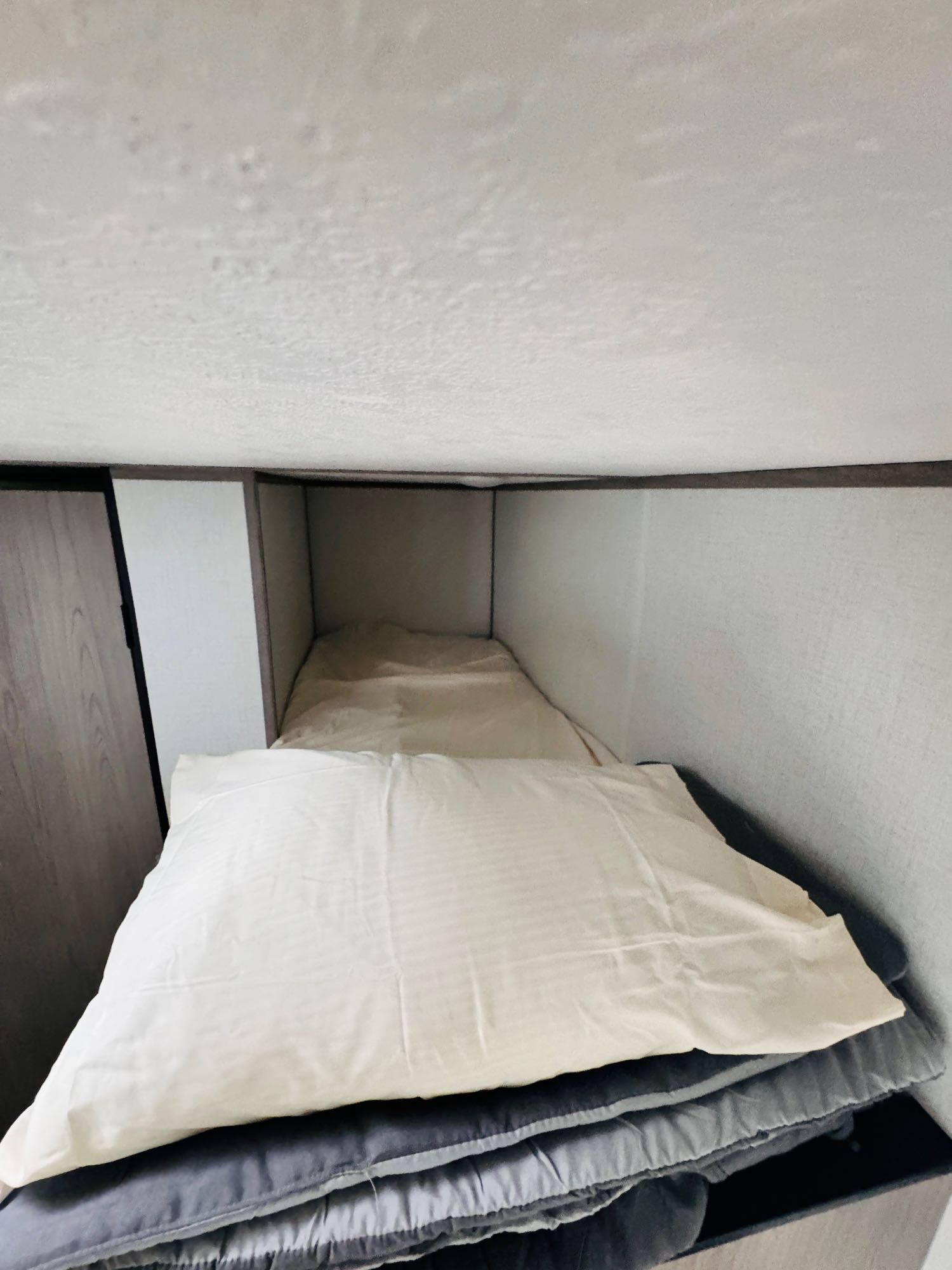 Inside an RV rental: a compact, lower bunk bed with a pillow and neatly stacked grey bedding.. Dutchmen 4ydtcmg18r8932632 2024