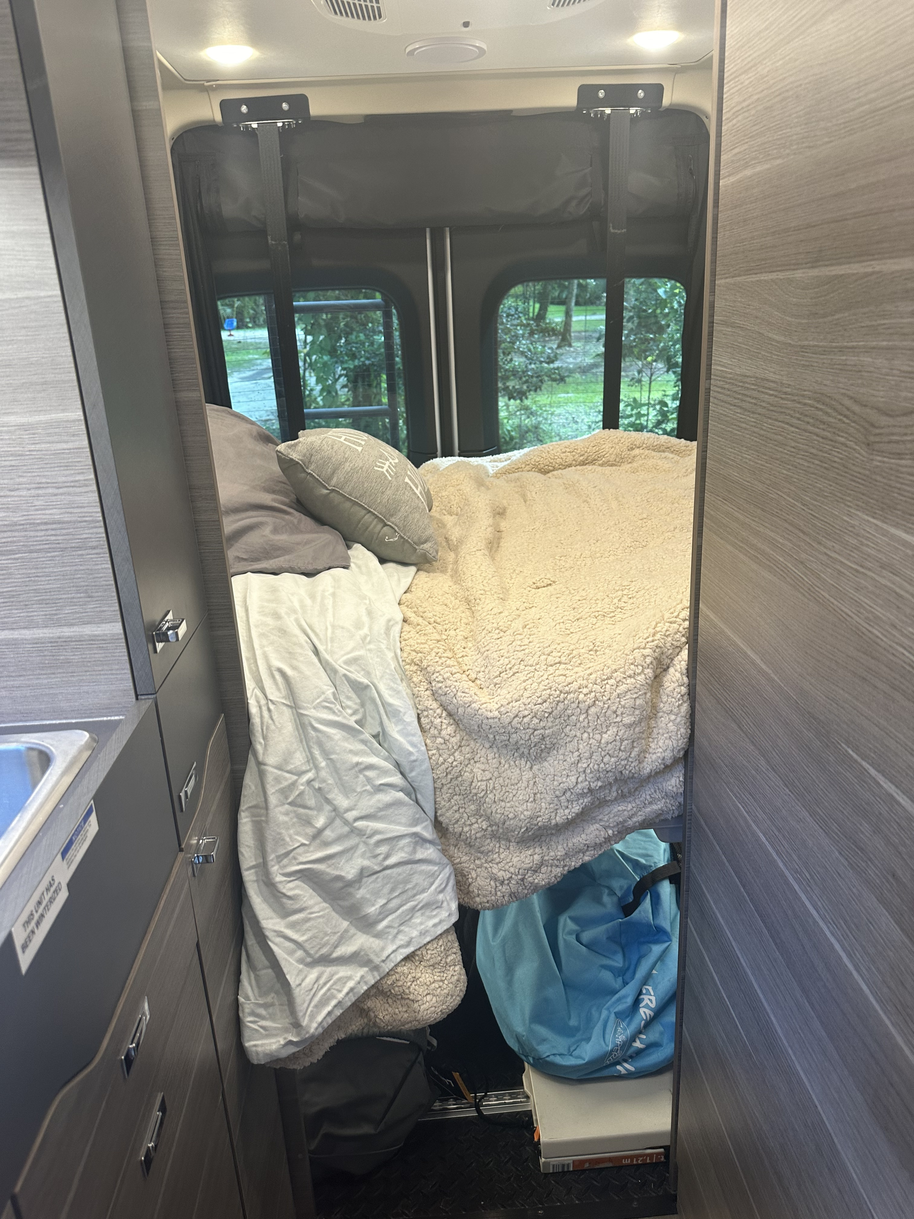 Inside this RV rental: a cozy bed with a fuzzy blanket, cabinets with a sink, and rear windows with a nature view.. Mercedes-Benz Jayco Terrain 2023