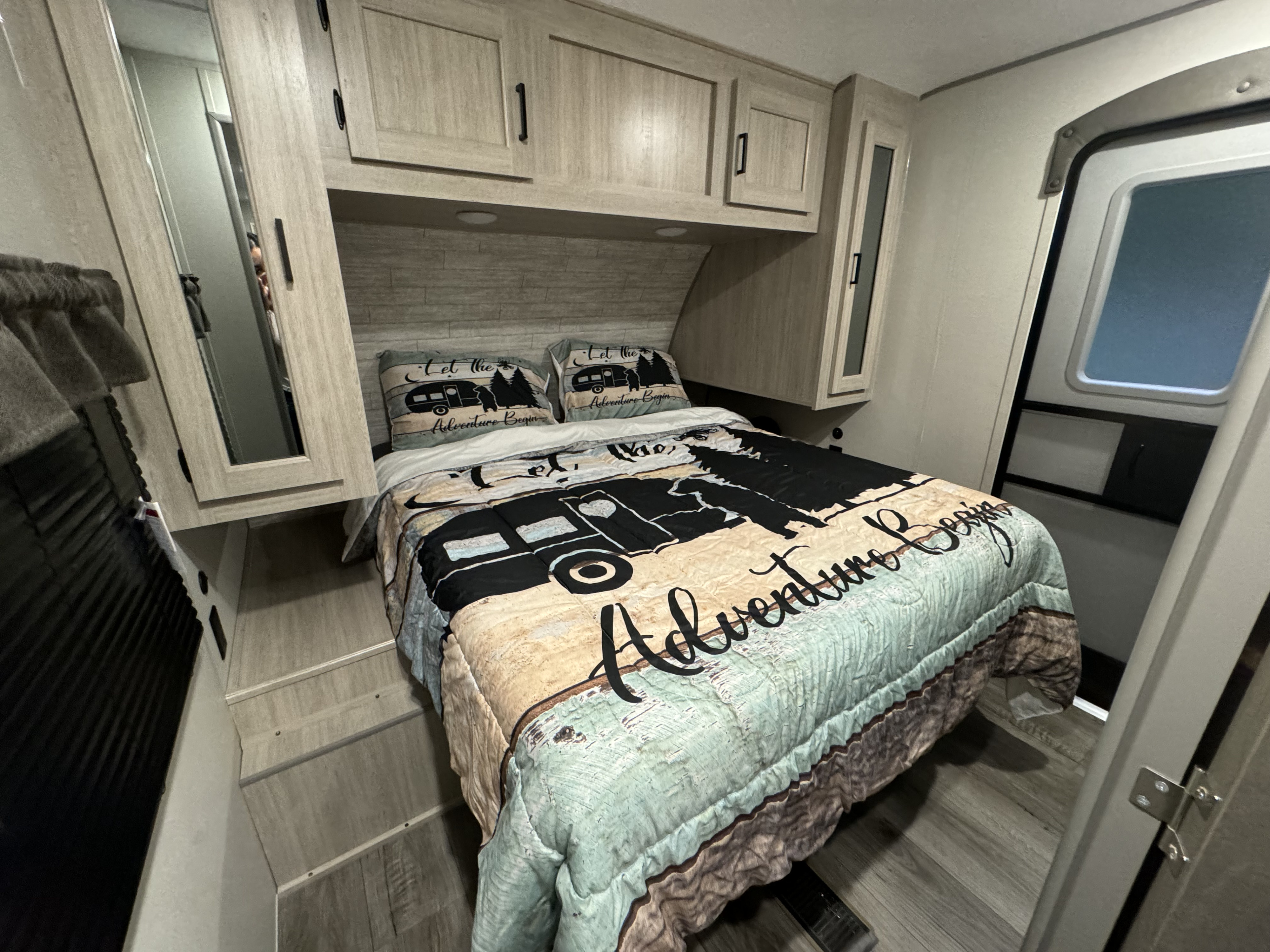 Cozy RV rental bedroom with adventure-themed bedding, light wood cabinets, and an entrance door. Perfect for travel!. Coachmen Catalina legacy edition 2024