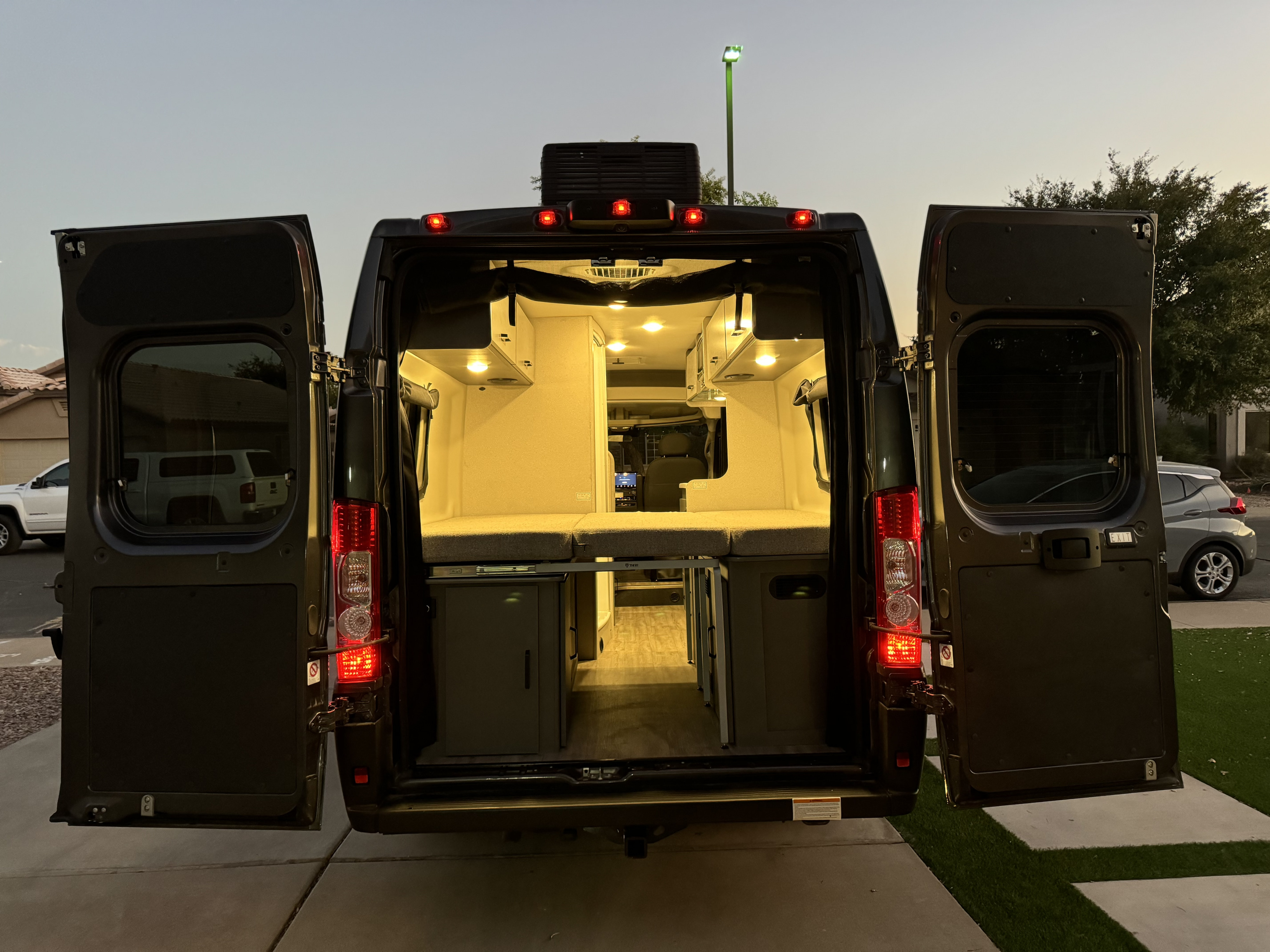 Explore a modern camper van RV rental interior at dusk: illuminated bed, cabinets, and amenities through open rear doors.. Thor Dazzle 2024