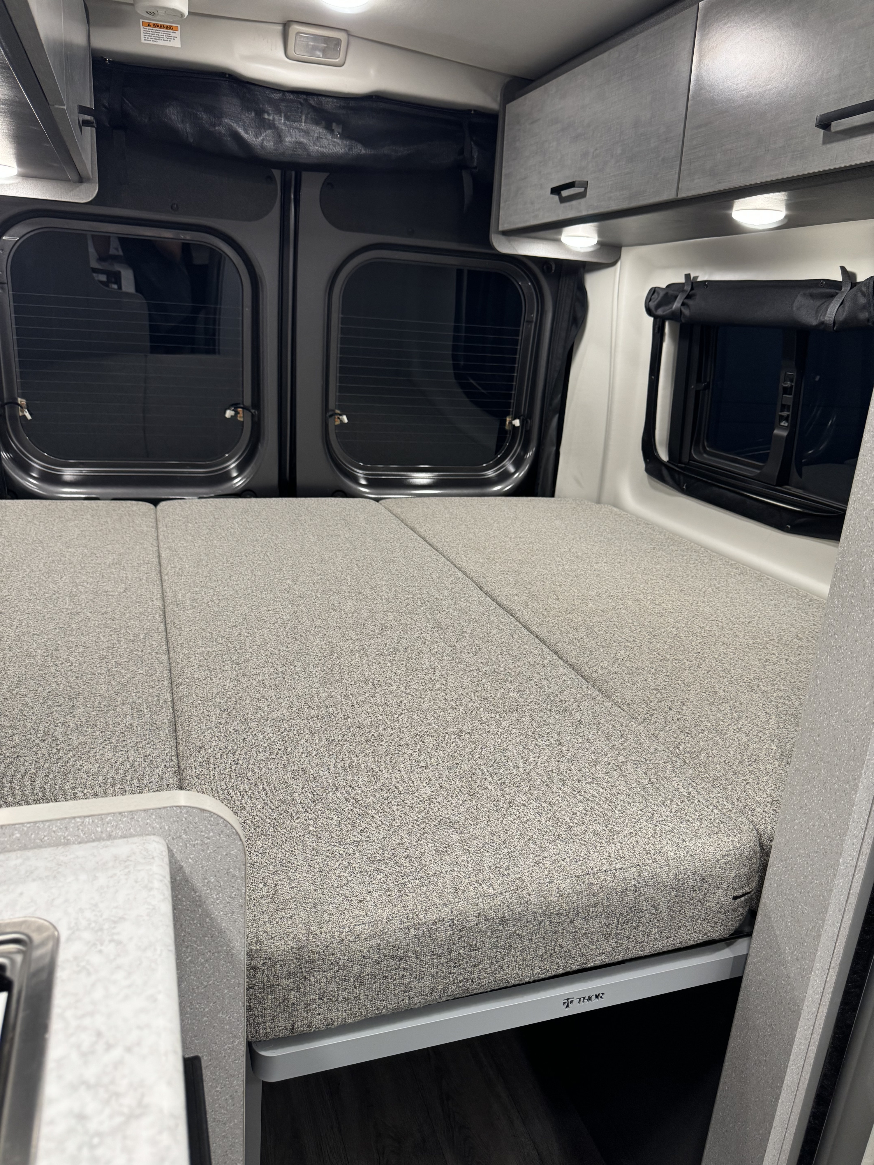 Comfortable RV rental interior with a gray convertible bed, ample overhead storage, and windows, ideal for travel.. Thor Dazzle 2024
