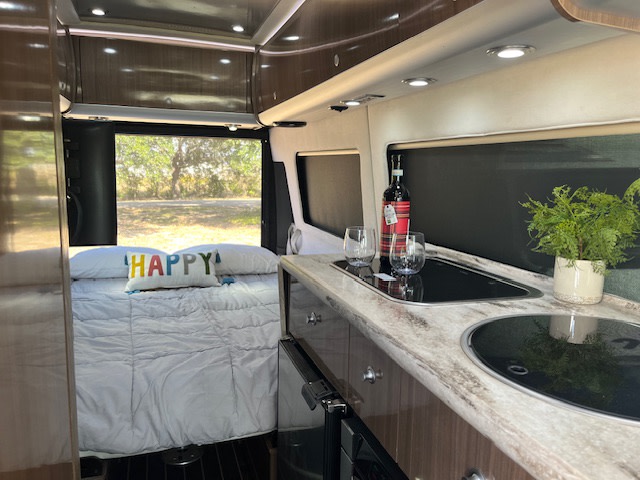 Cozy rv rental interior: bed with "HAPPY" pillow, kitchen counter with wine & plant. Your adventure awaits!. Airstream Interstate 2018