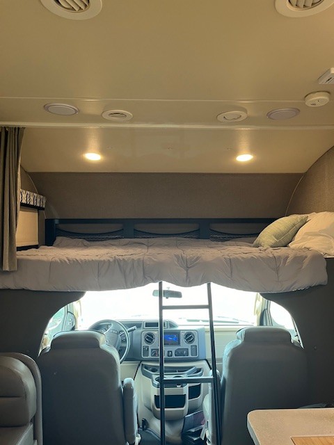 View from inside an RV rental, showing the front cockpit area and a comfortable over-cab bunk bed with a ladder.. Jayco Greyhawk 2020