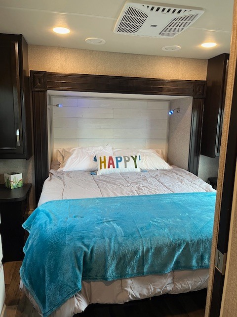 Cozy RV rental bedroom features a comfy bed with a "HAPPY" pillow, overhead AC, and bedside storage.. Jayco Greyhawk 2020