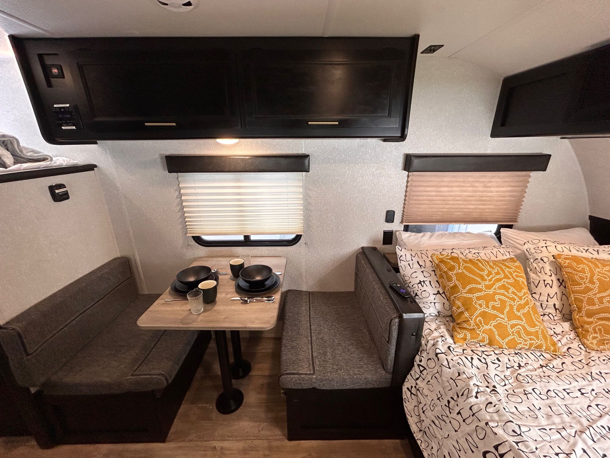 Interior of an RV rental showing a dinette with bench seating and set table, alongside a cozy bed with patterned linens, and overhead cabinets.. Forest River Cherokee Wolf Pup 2021