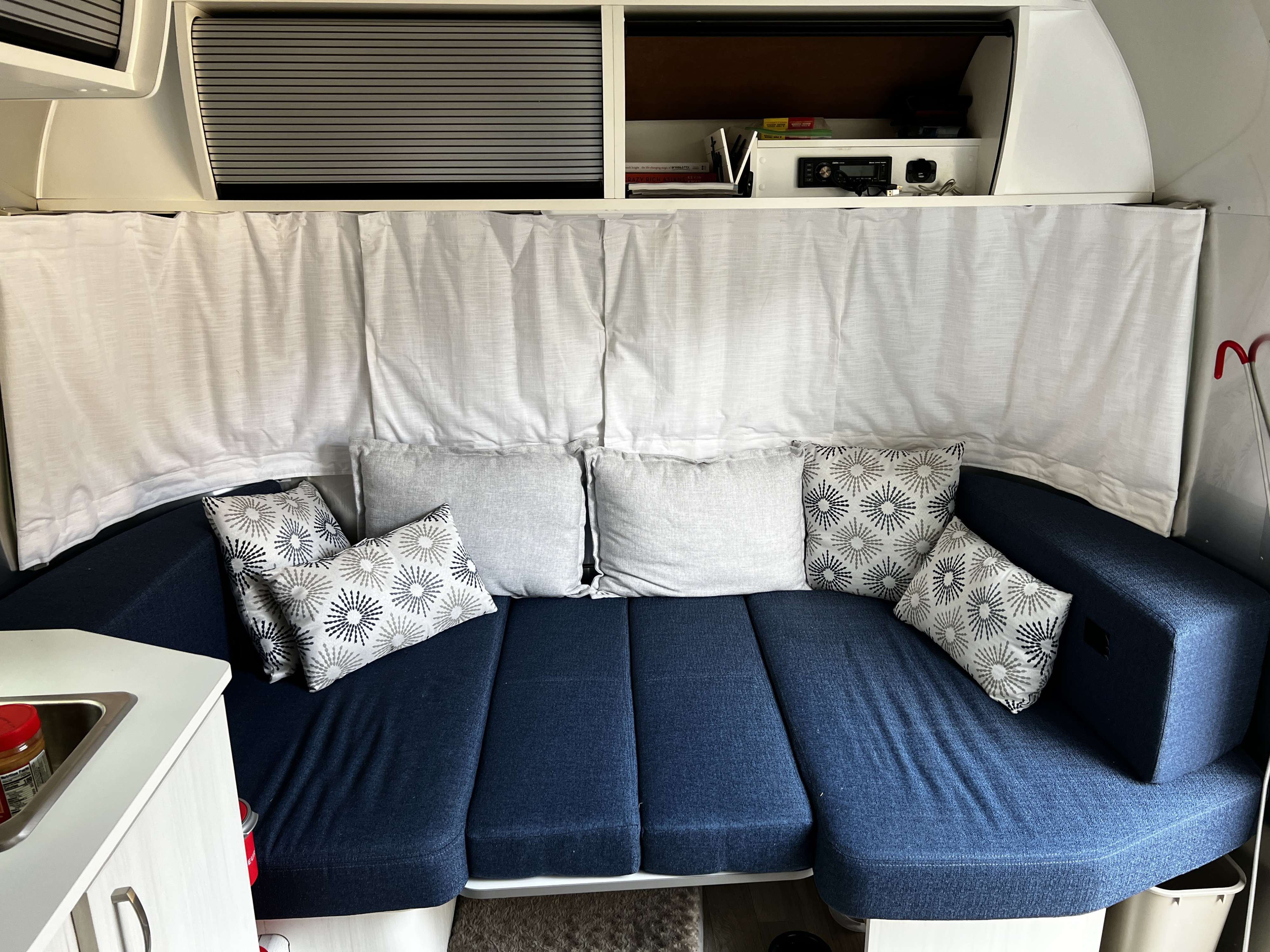 RV rental interior: blue sofa with patterned and plain pillows, practical overhead storage, and white privacy curtains.. Airstream Bambi 2020