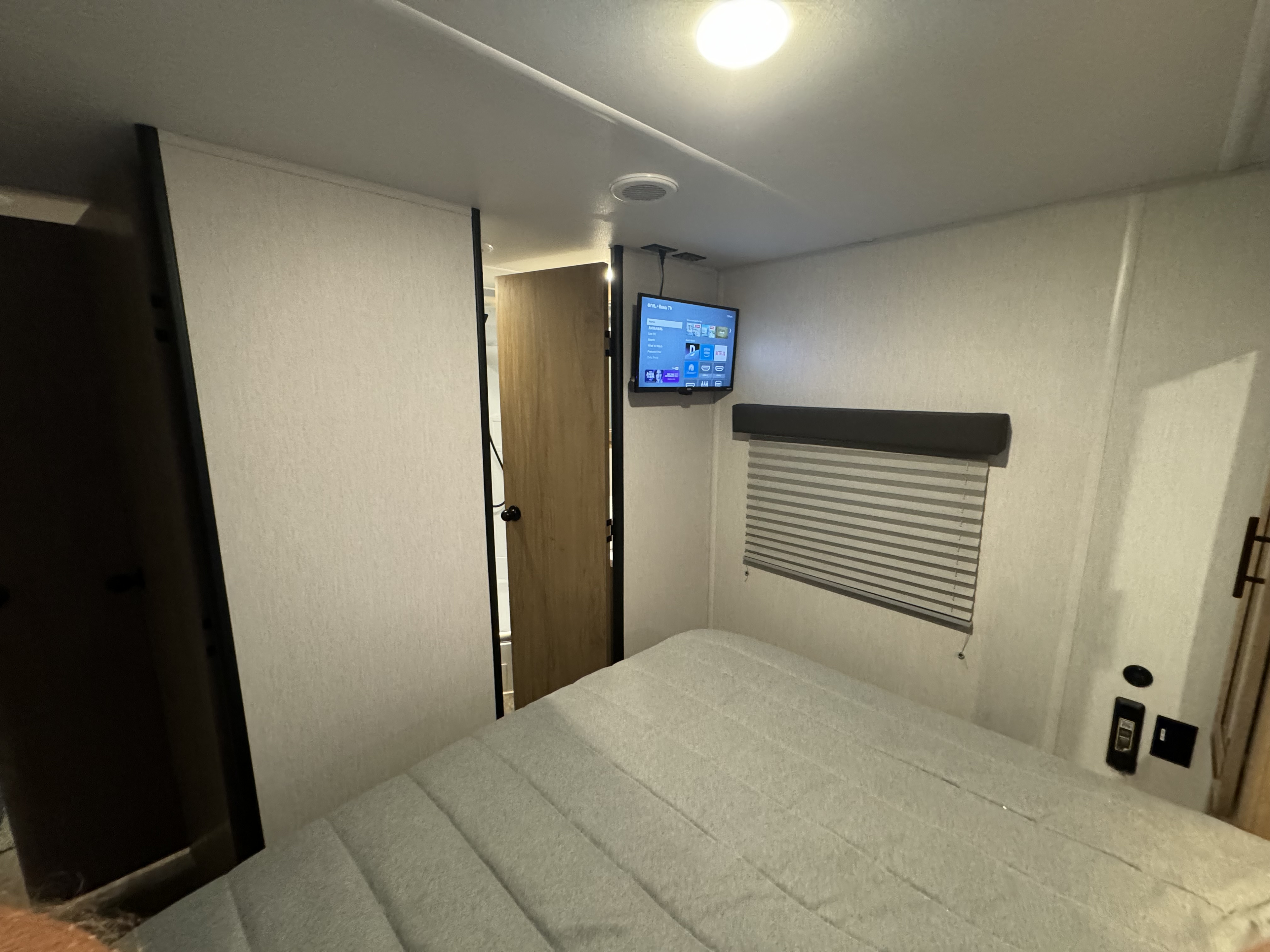 Inside an RV rental: a compact bedroom with a soft bed, mounted smart TV, window blinds, and ceiling light.. Forest River Cherokee Grey Wolf 2024
