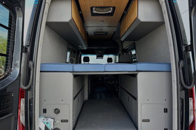 Experience an RV rental: a cozy camper van interior with a raised blue bed, overhead cabinets, and large rear storage.. Mercedes-Benz Sprinter 2020