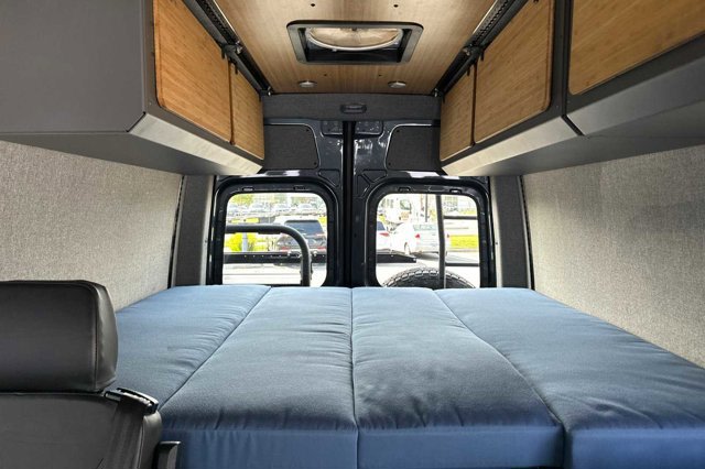 RV rental van interior with comfy bed, wood overhead storage, and rear windows looking out.. Mercedes-Benz Sprinter 2020