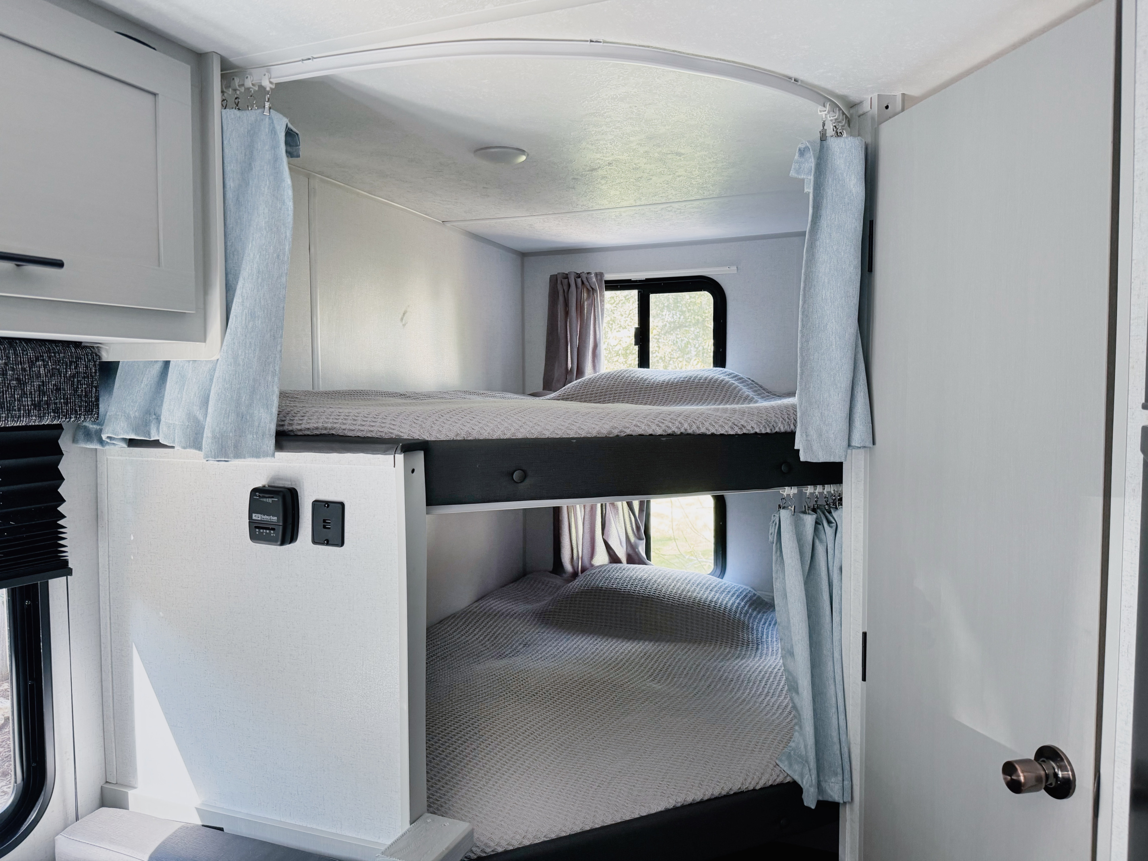Inside an RV rental: Brightly lit bunk beds with light gray covers, blue privacy curtains, and windows offering outdoor views.. Forest River Wildwood FSX Platinum 2021