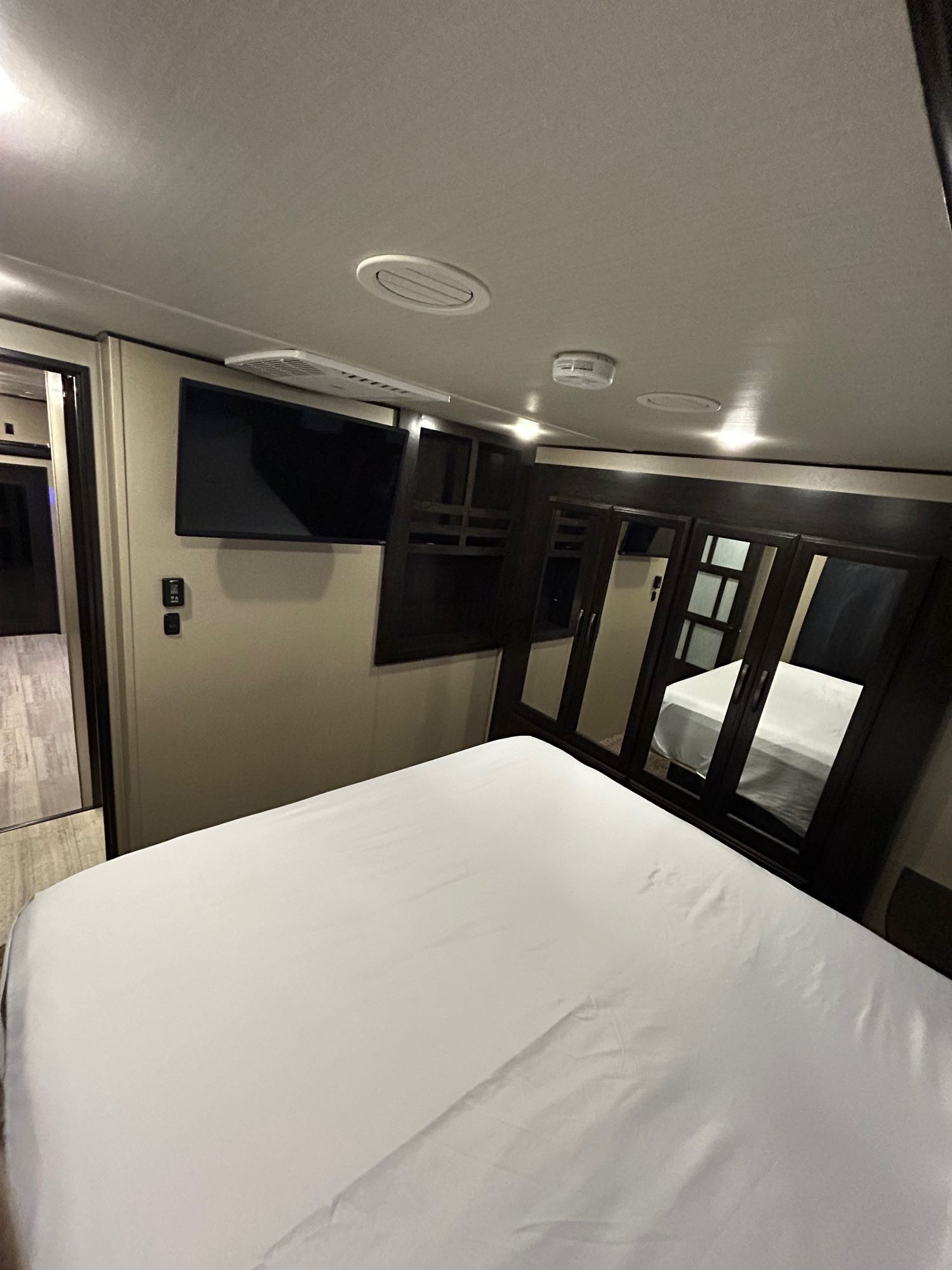 This RV rental bedroom features a white bed, wall-mounted TV, and dark mirrored wardrobe, with a doorway visible left.. Grand Design Momentum 2017