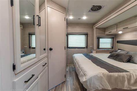 Relax in this RV rental's cozy bedroom, featuring a large bed with a grey throw, two windows, overhead cabinets, and a mirrored wardrobe.. Thor A.C.E. 2020