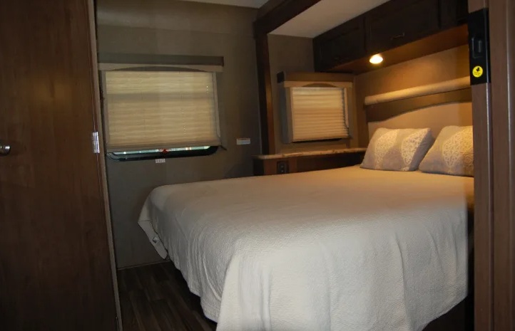 This cozy RV rental bedroom features a large bed, two windows with blinds, overhead storage, and warm lighting.. Thor A.C.E. 2020