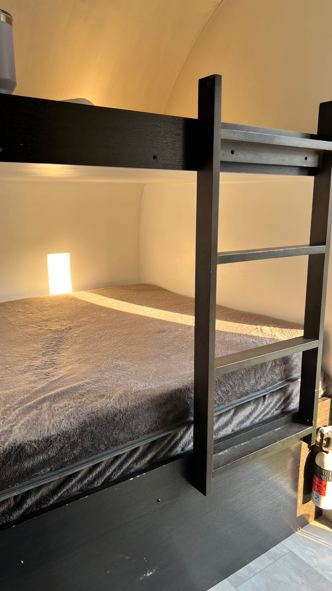 Cozy RV rental interior: lower bunk bed with a soft grey blanket, sunlight streaming in. A fire extinguisher is visible.. Braxton Creek Free Solo FAM 2021