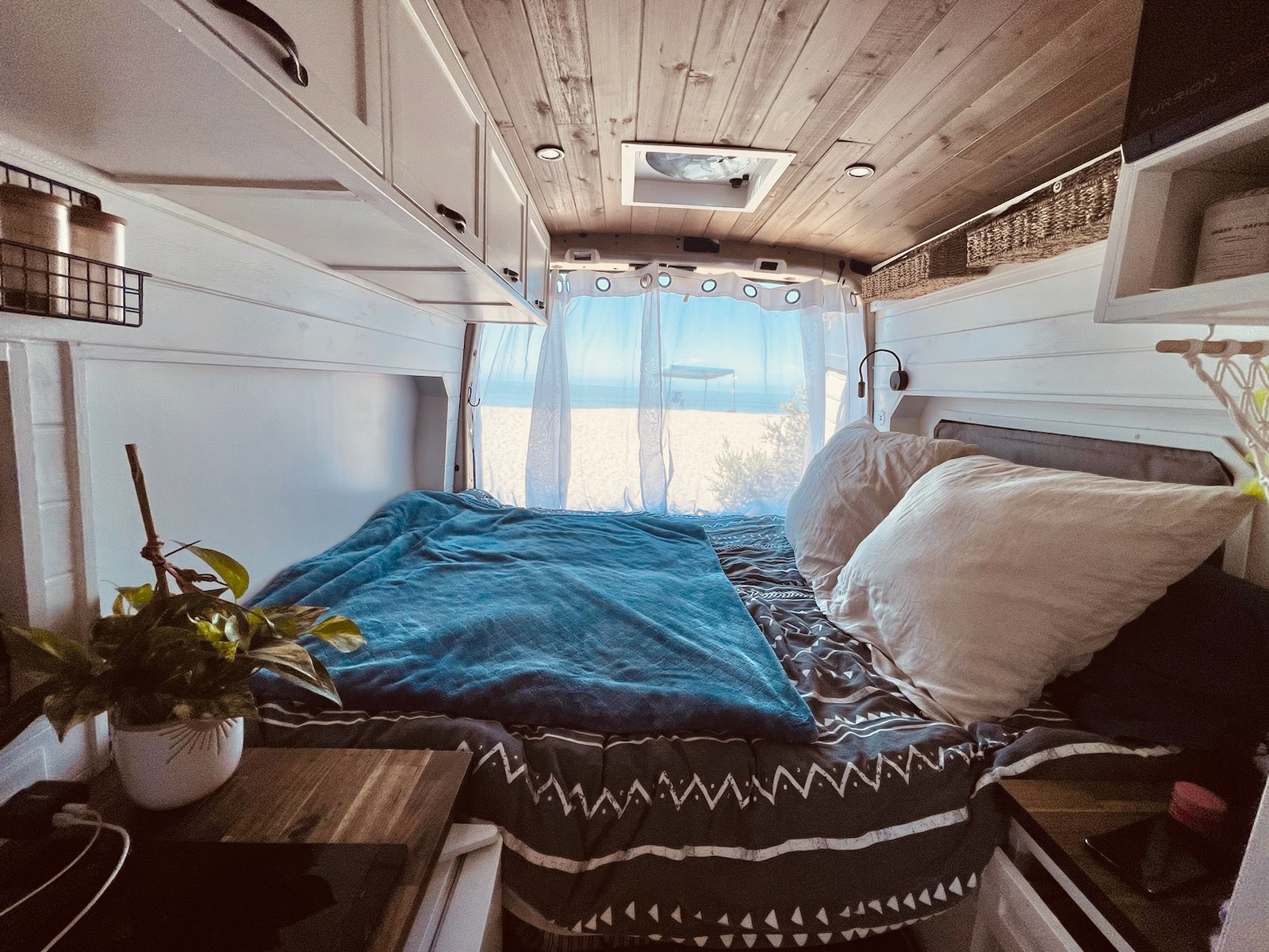 Experience comfort in this RV rental bedroom with a serene beach backdrop. Wooden ceiling, ample storage, and warm lighting.. Ford Transit 2020