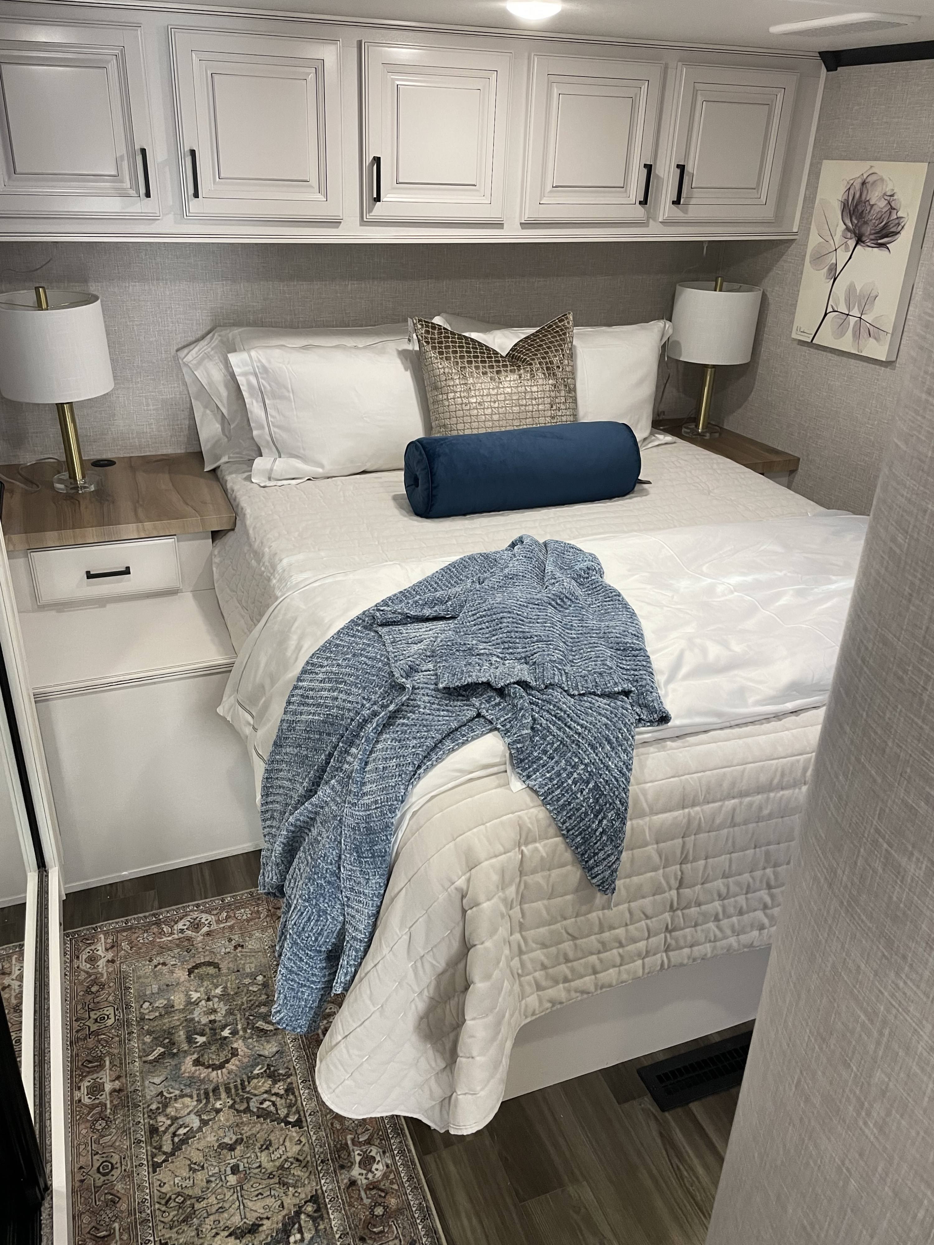 Cozy RV rental bedroom with white bedding, blue accents, overhead cabinets, lamps, and a patterned rug.. Highland Ridge RV Open Range 2024