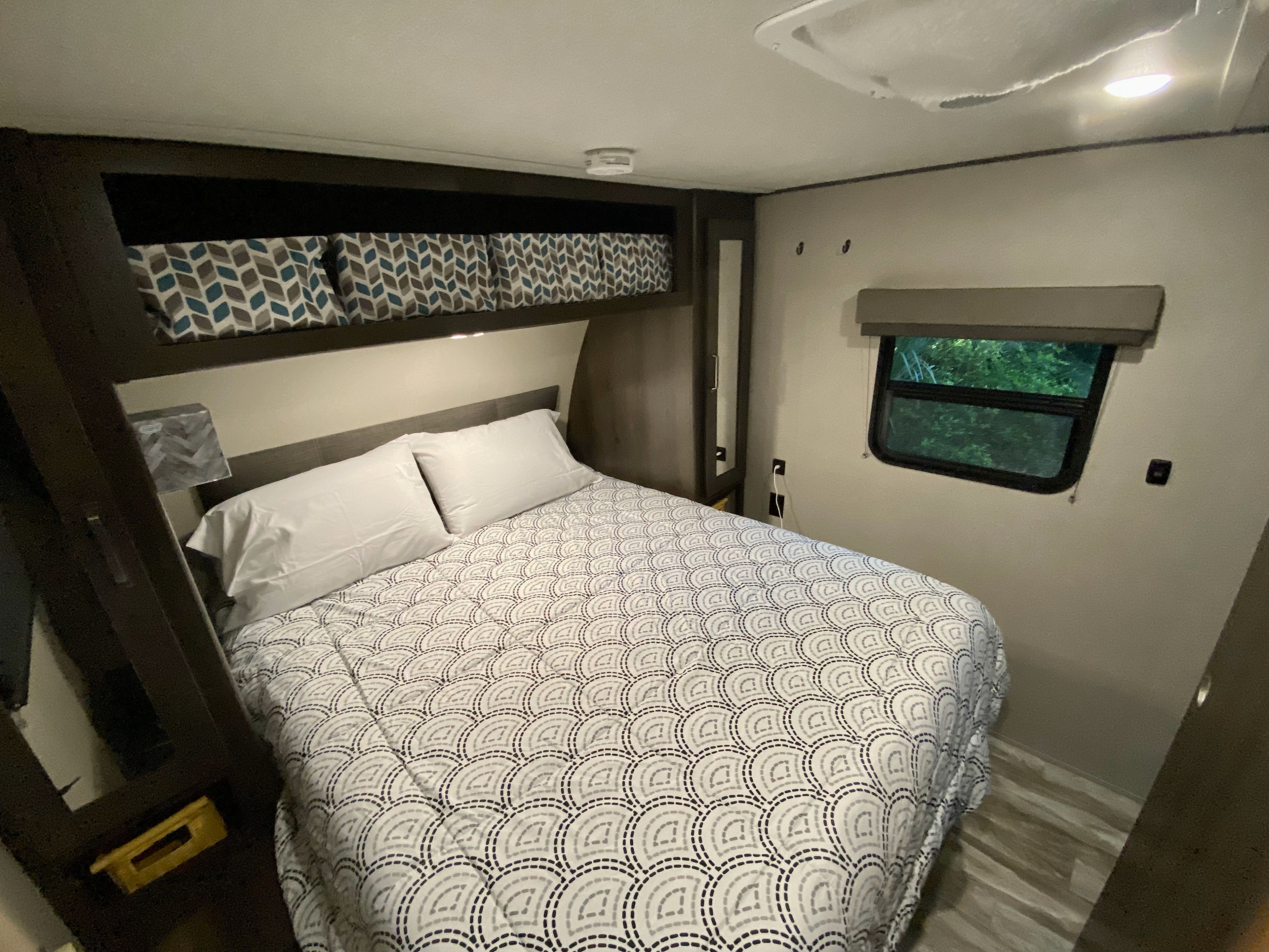 Cozy RV rental bedroom with white pillows, patterned comforter, overhead storage, and a window view.. Grand Design Transcend Xplor 2021