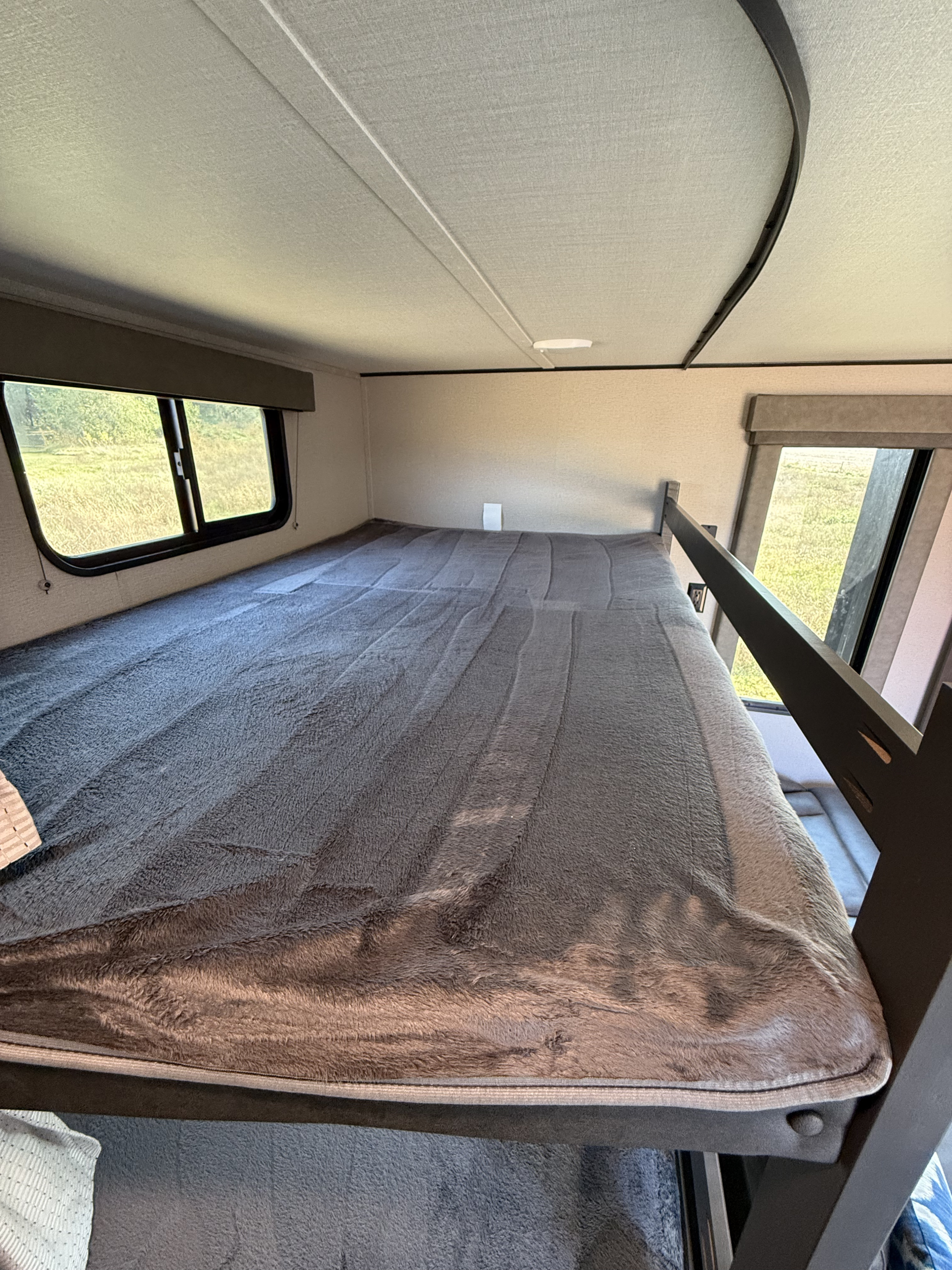 Cozy top bunk bed in an RV rental, featuring a soft blanket and bright window overlooking a field.. Grand Design Transcend Xplor 2021