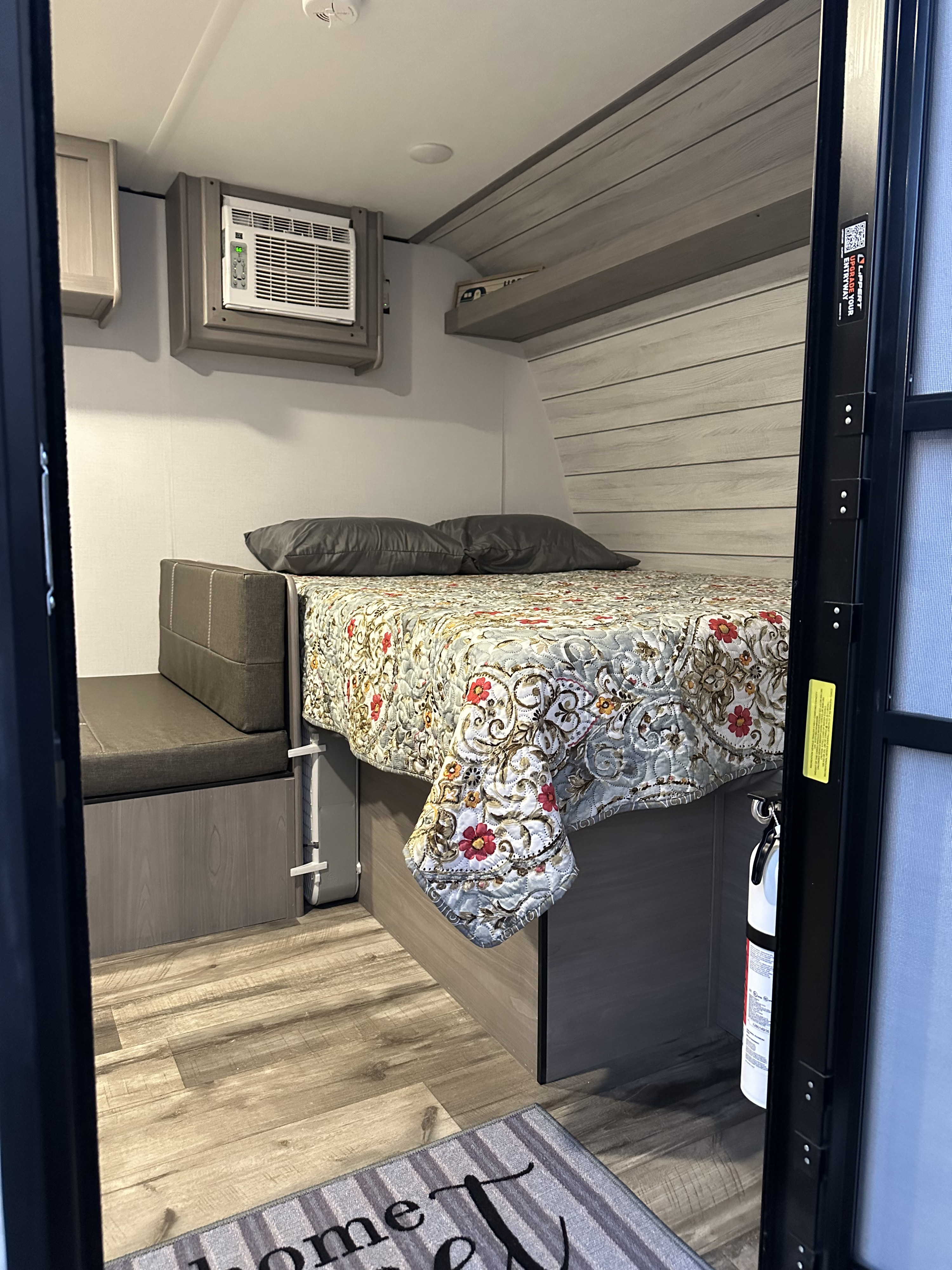 Cozy RV rental interior: A compact bedroom with a floral-quilted bed, bench seating, wall-mounted AC, and wood-grain floor.. Coleman Lantern 2024