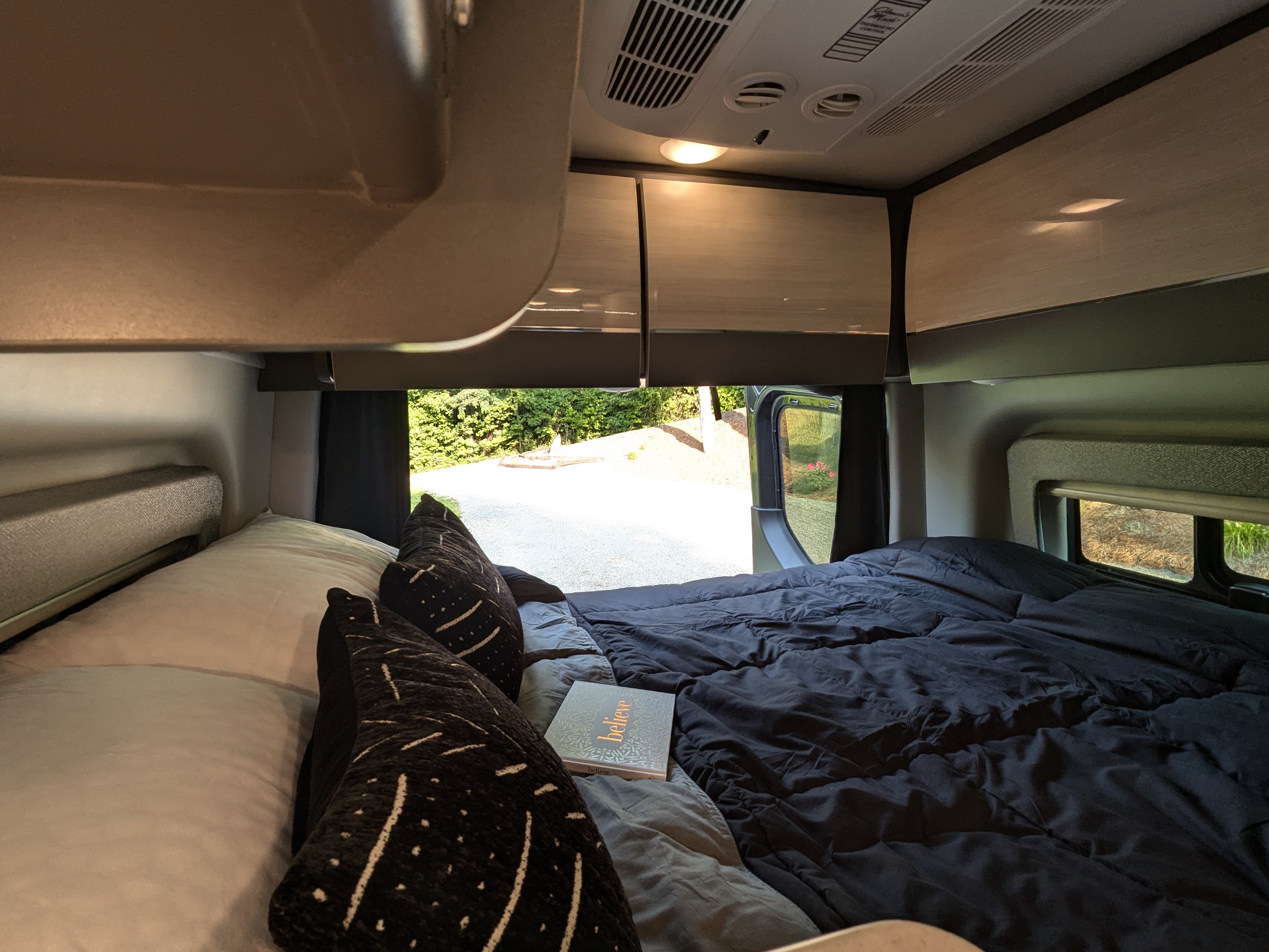 Cozy rv rental bed area with pillows, a book, overhead cabinets & vents, offering an inviting view of nature from the open rear door.. Thor Motor Coach Sanctuary 19L 2024