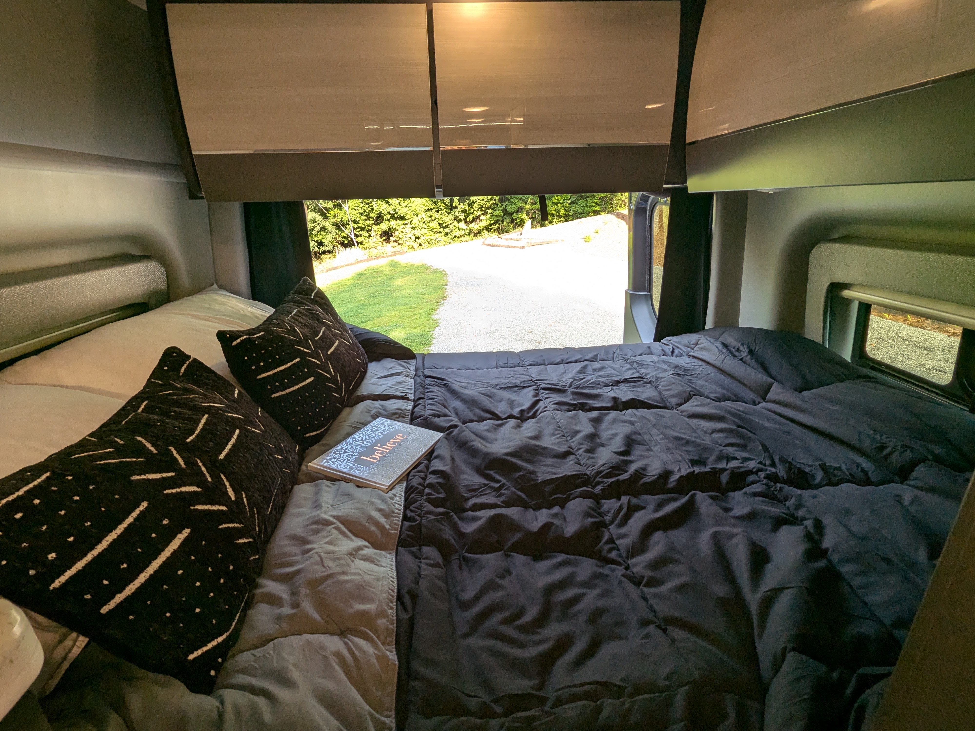 Cozy RV rental bedroom featuring a comfy bed with patterned pillows and a "believe" book, offering a peaceful outdoor view.. Thor Motor Coach Sanctuary 19L 2024