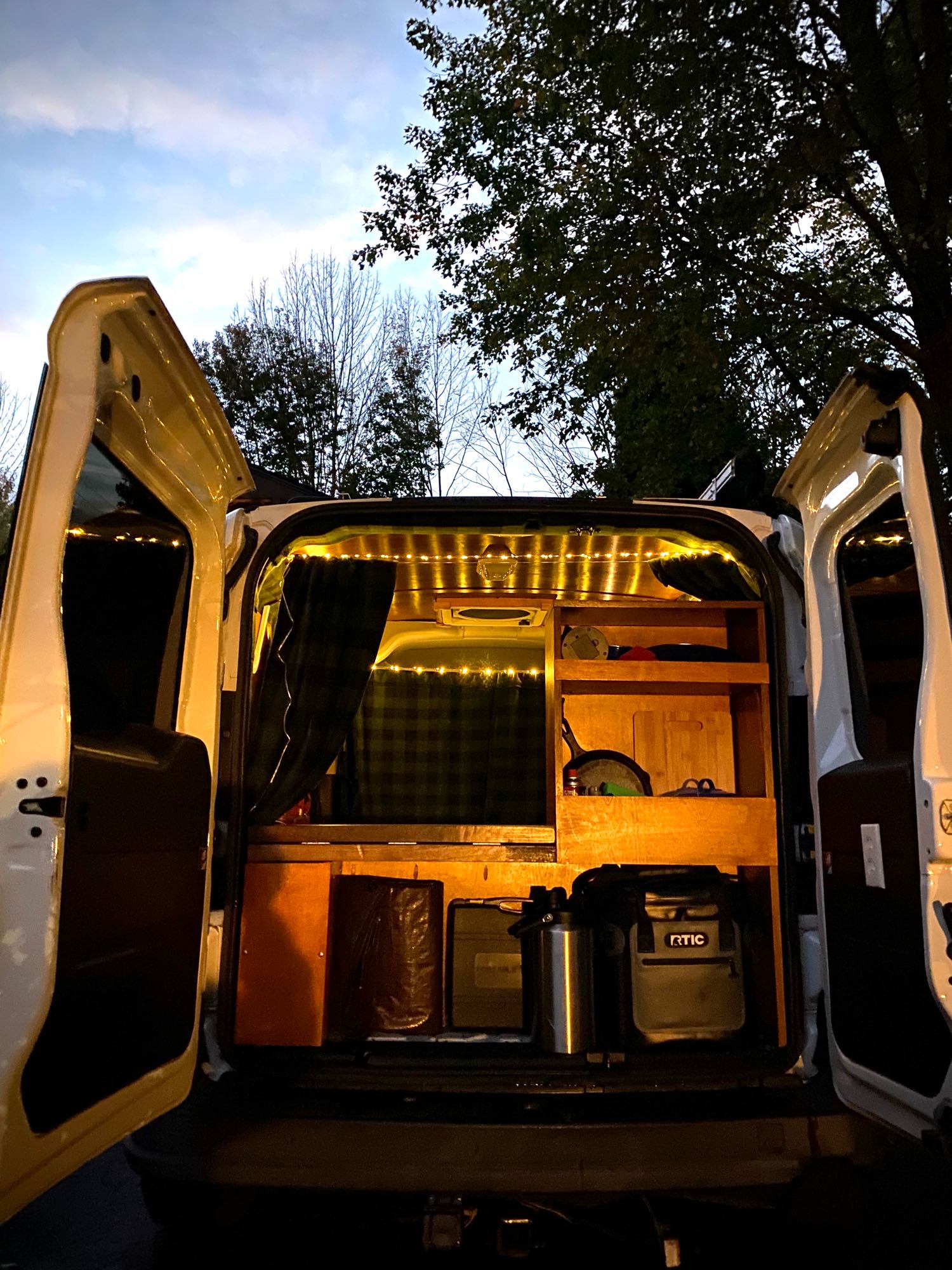 Cozy camper van interior seen from the rear, doors open. Custom wood build, warm string lights, & storage. Perfect RV rental.. Dodge Ram Promaster City 2015