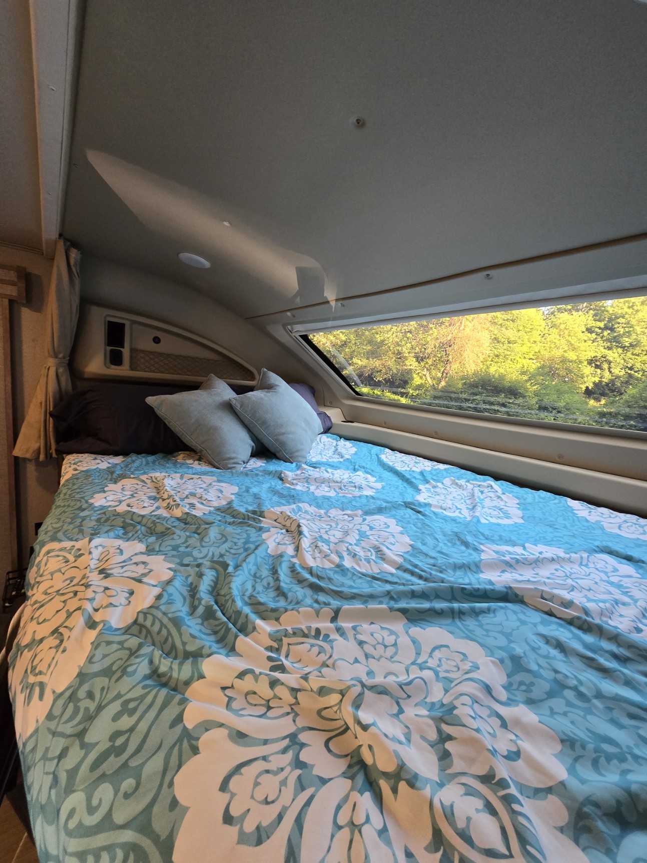 Cozy RV rental bedroom features a comfy bed with blue floral duvet and large window looking out at lush green trees.. Entegra Coach Odyssey 2020