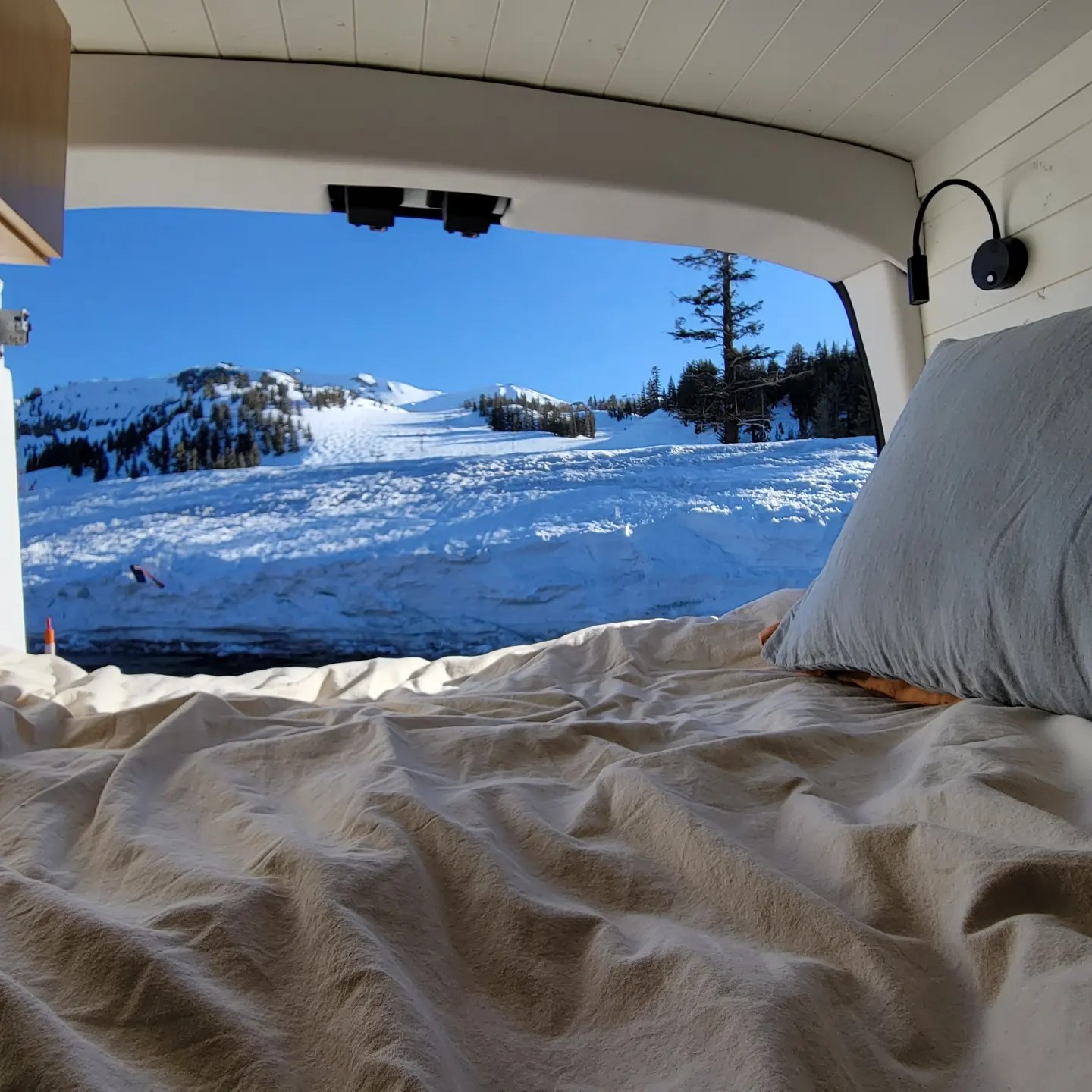 Inside an RV rental: a cozy bed overlooking snowy mountains, ski tracks, and a clear blue sky. Winter adventure awaits!. Ford Transit 2020