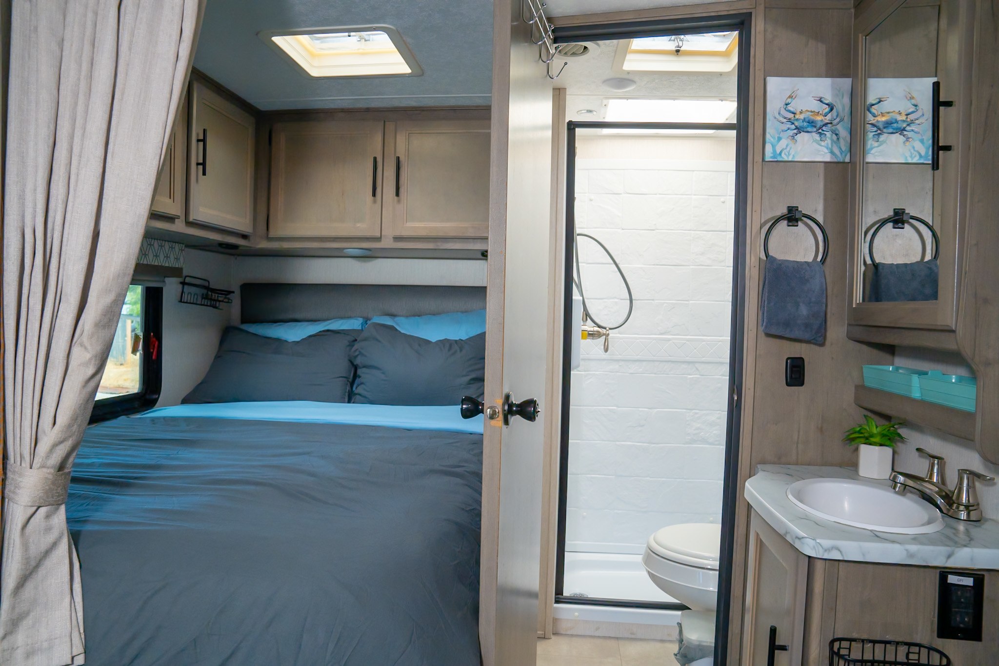 Compact RV rental interior featuring a cozy bedroom area with gray and blue bedding, and a functional bathroom with shower and vanity.. Coachmen Prism 2020