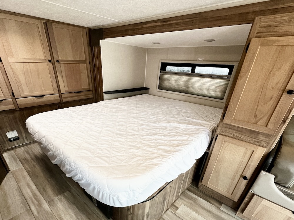 Spacious RV rental bedroom featuring a large bed with a white quilted cover, built-in wooden storage, and a window with blinds.. Coachmen Prism 24FS 2024