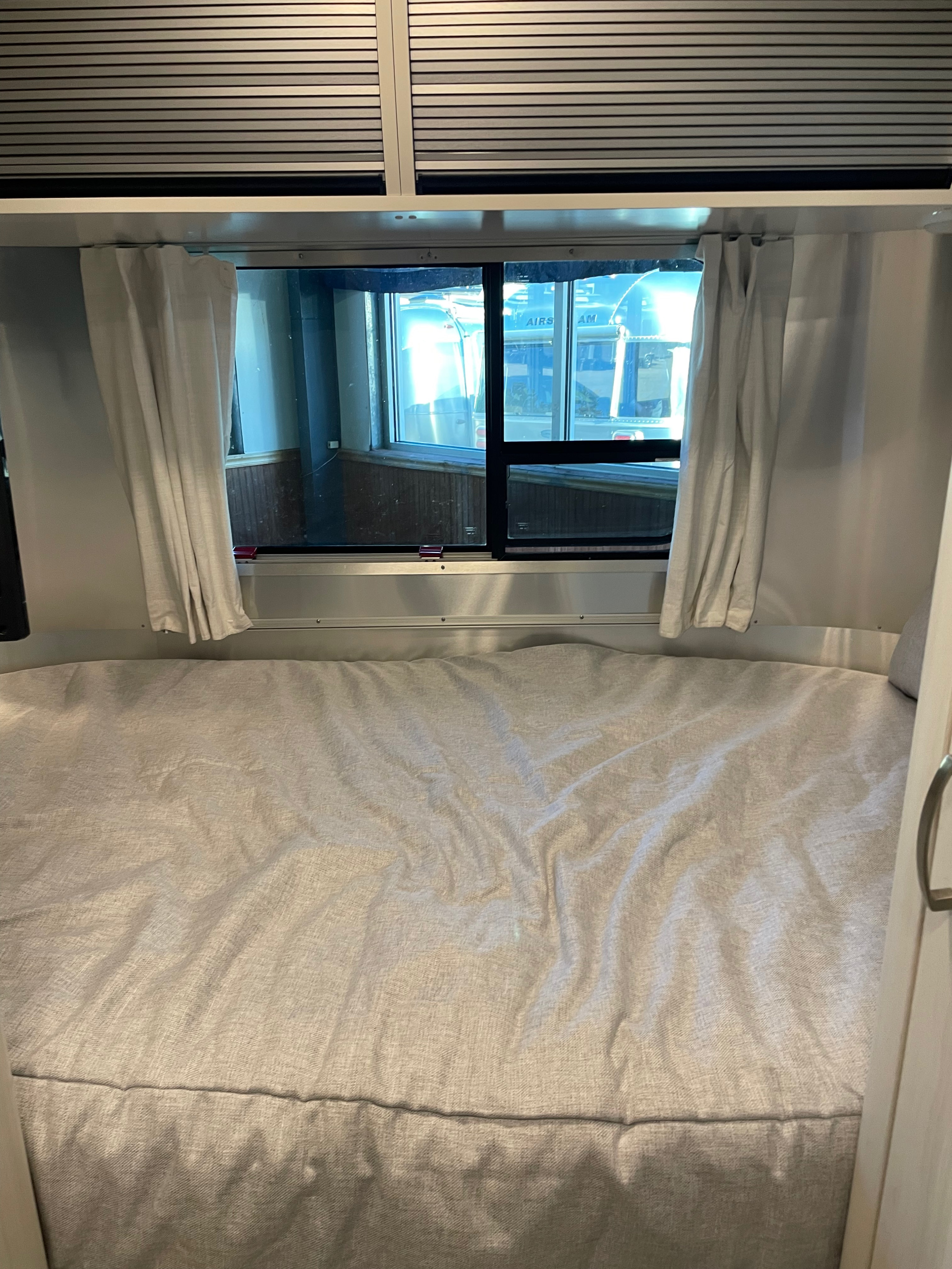 RV rental interior: cozy bed with open curtains, revealing Airstream trailers outside a large window. Overhead blinds are closed.. Airstream Bambi 2022