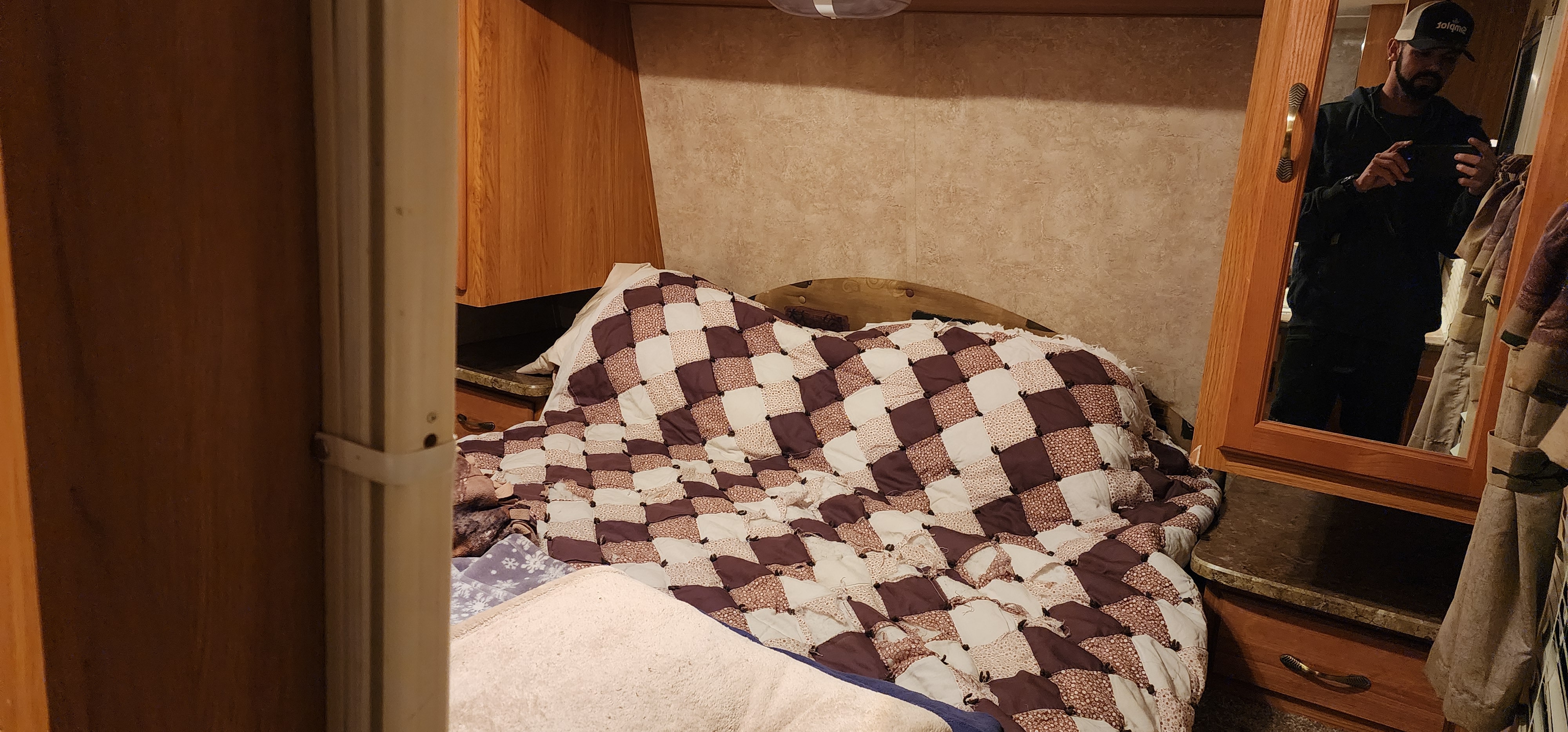 Cozy RV rental bedroom featuring a checked quilt, wood cabinets, and a man's reflection in the mirror.. Keystone Springdale 2010
