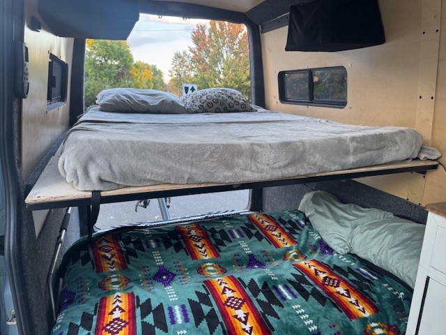 RV rental interior featuring custom-built bunk beds. Top bunk has gray bedding, bottom bunk has a colorful patterned blanket.. Mercedes-Benz Sprinter 2021