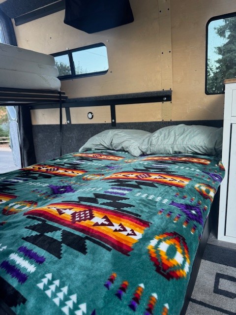 Colorful bed with Southwestern blanket in an RV rental interior, showing upper bunk & windows.. Mercedes-Benz Sprinter 2021